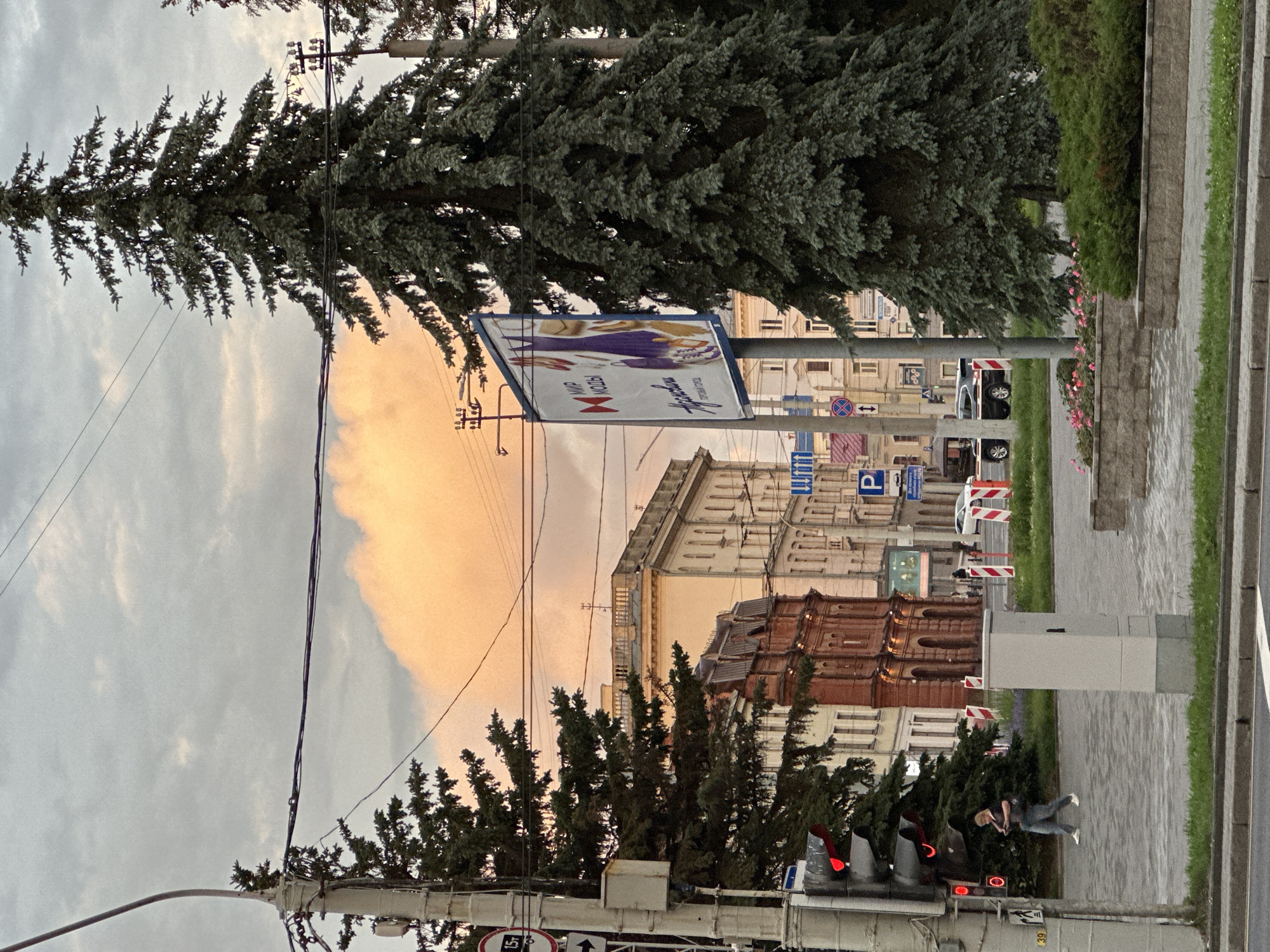 The image depicts a street scene in Minsk, Belarus, with a large evergreen tree dominating the foreground on the right. Behind the tree, a billboard advertising "МИР МОДЫ" (World of Fashion) is visible. The background features several buildings, including a red brick structure and a lighter-colored, multi-story building. Overhead wires and a traffic light are also present. On the left side of the image, a woman walks along a paved sidewalk. The sky is overcast with a large, orange-tinted cloud. The overall impression is an urban environment during what appears to be either dawn or dusk, given the lighting conditions and the color of the cloud.