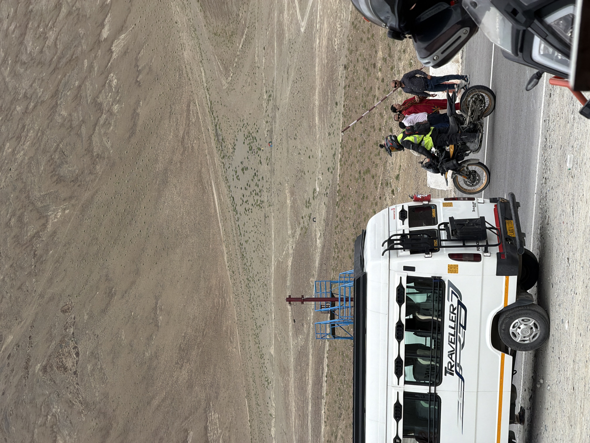 The image shows a group of people and vehicles on a road with a vast, arid mountain landscape in the background, indicative of a location like Chumkar, India. In the foreground, a white van labeled "TRAVELLER" is parked. To the right of the van, a person on a motorcycle wearing a high-visibility yellow jacket is stopped near a barrier. Several other individuals are standing near the barrier, appearing to interact. The landscape consists of barren, dusty hills with sparse vegetation. A blue metal structure with railings is visible on top of the van. The sky is overcast.