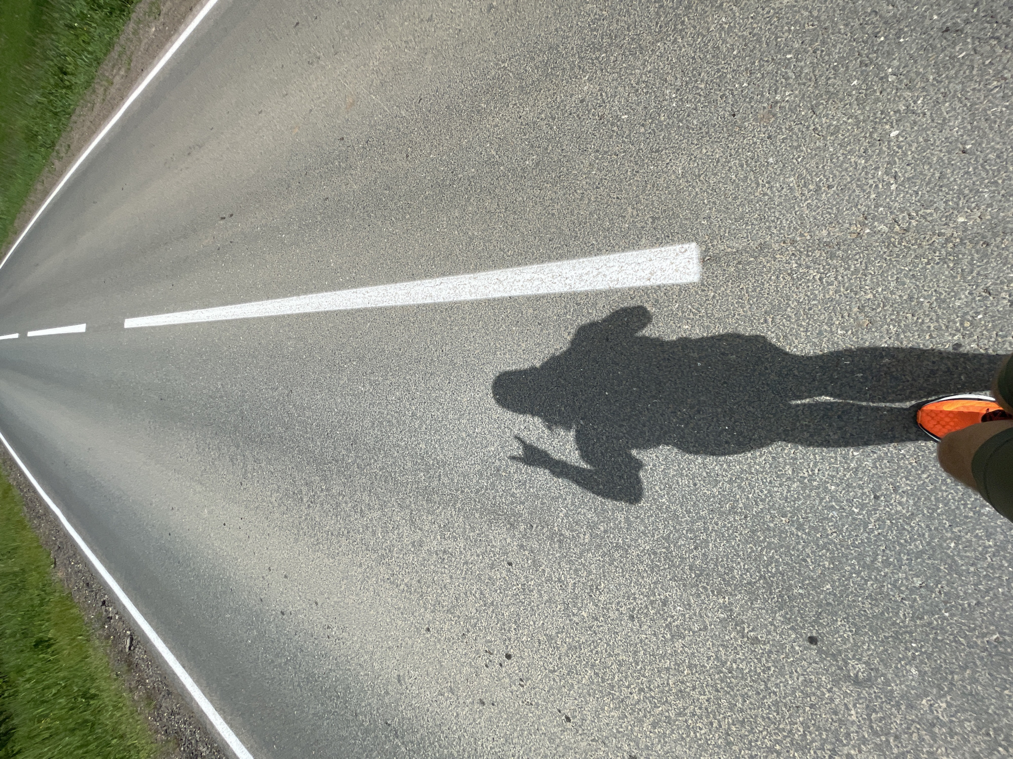 The image presents a high-angle, top-down view of a person's shadow and foot on an asphalt road. A distinct, elongated shadow of a person, likely the photographer, occupies the midground and foreground, extending from the lower right toward the center. The shadow's left arm is raised, with two fingers clearly extended, resembling a peace sign. The person's lower right leg, clad in dark shorts, and a brightly colored orange athletic shoe are visible at the bottom right corner of the frame.
The road is made of dark grey asphalt with a coarse, textured surface. A solid white line marks the right edge of the paved road, bordering a strip of dirt and green grass. Further into the road, a dashed white line, likely indicating a lane division or center line, is visible in the upper left quadrant. The strong, well-defined shadow suggests bright, direct sunlight and a high sun angle, indicative of midday conditions. The setting appears to be a rural or suburban road with natural vegetation along its edge. There is no visible text in the image. The scene captures a moment during a walk or run on a clear, sunny day.