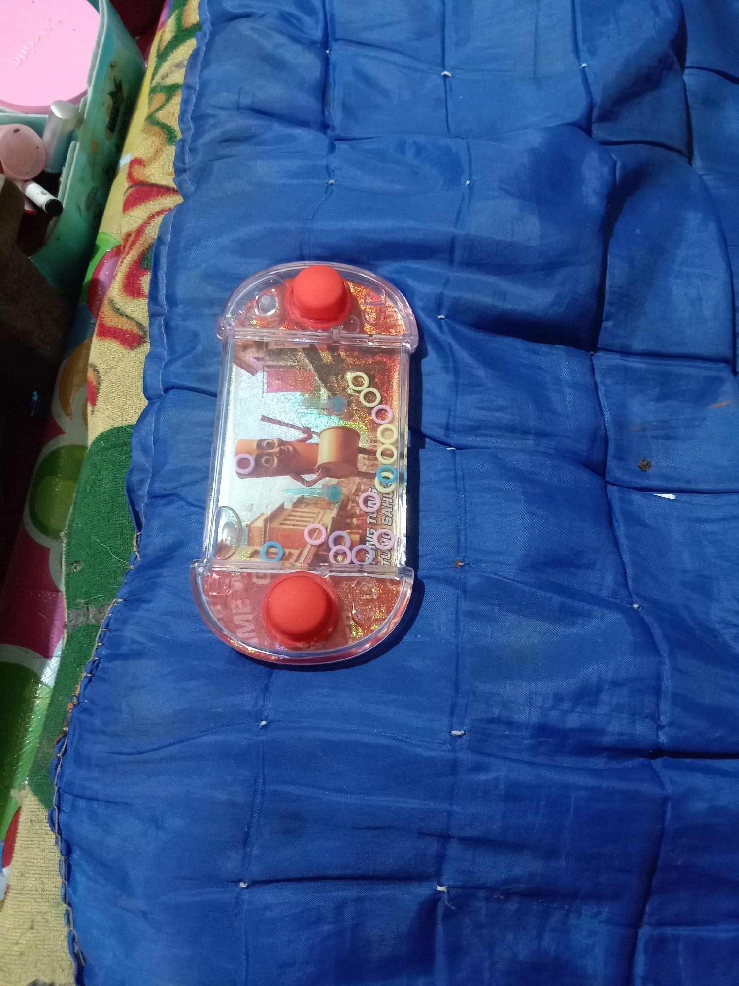 An overhead shot shows a vintage water game toy on a blue quilted surface. The toy is rectangular with rounded ends and contains a colorful insert depicting cartoon characters and rings. Two red buttons are visible, one at the top and one at the bottom of the toy. The blue surface has visible stitching and appears to be a mat or cushion. The background on the left shows a portion of a colorful patterned fabric. The image is taken indoors and does not provide any information about the city of Jumput, Indonesia.