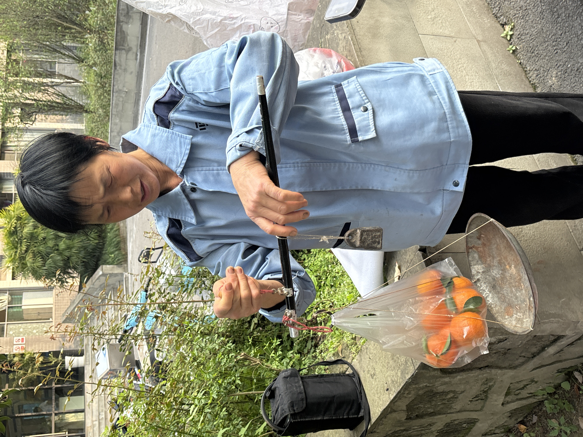 The image depicts an outdoor scene in Jiangjin, China. A female individual with short, dark hair is shown from the waist up, wearing a light blue collared jacket with dark blue shoulder panels and horizontal stripes on the chest pockets, buttoned cuffs, and black trousers. Her eyes are partially closed. She is holding and manipulating a traditional steelyard scale: her left hand grips the upper suspension point of the scale, while her right hand supports the main black bar. Suspended from the left side of the steelyard is a clear plastic bag containing approximately seven bright orange tangerines with green leaves, positioned above a worn, oval-shaped metal pan. A dark, rectangular metal counterweight hangs from the right side of the steelyard. To the left of the individual, a black fabric shoulder bag rests on a low stone-faced wall. The ground beneath the subject is paved. The background features dense green foliage and small plants, behind which multi-story buildings with visible windows, balconies, and stone steps are present, indicating an urban or semi-urban environment.
