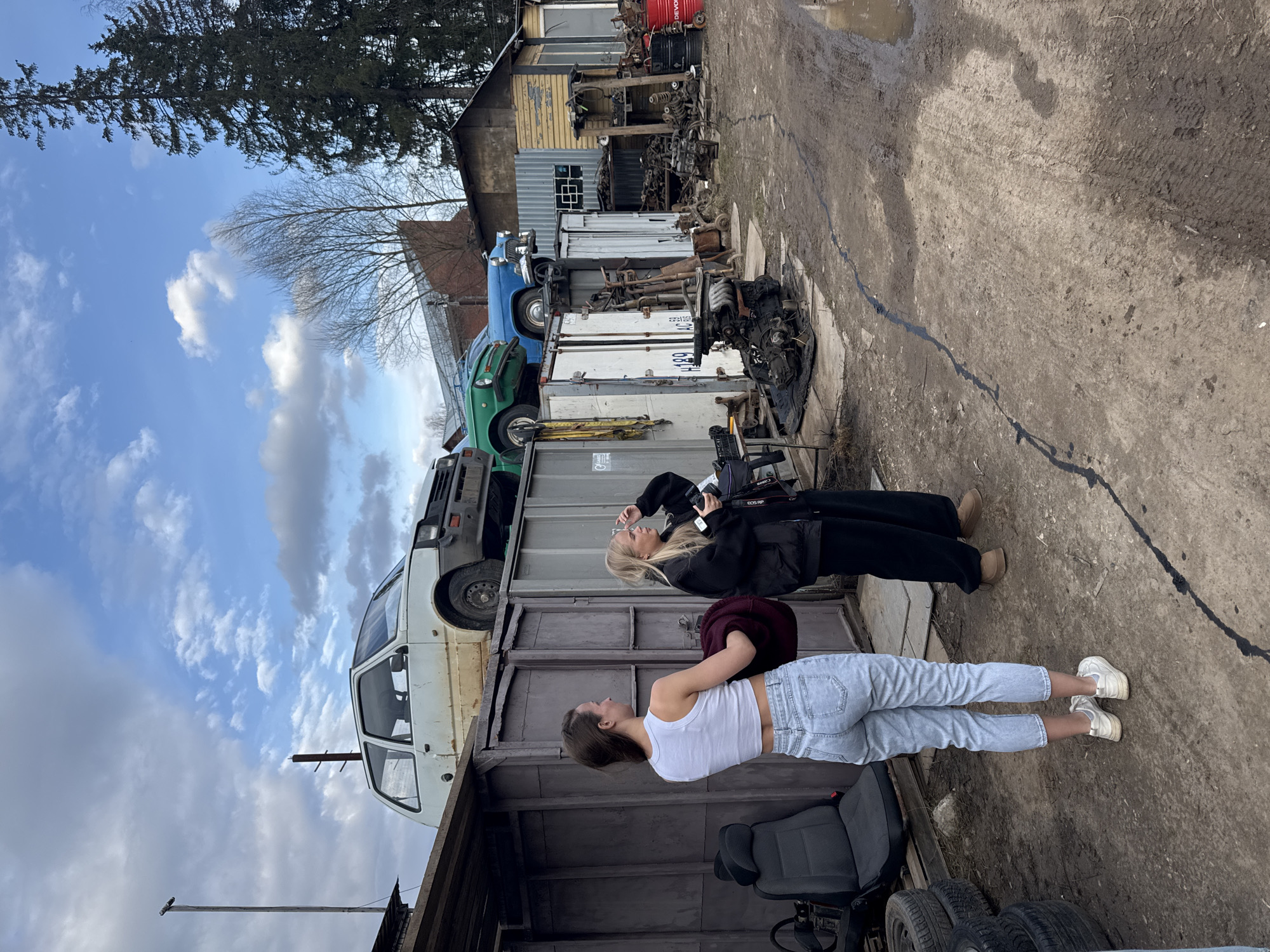 Two young women are depicted in an outdoor auto junkyard setting in Chaikovsky, Russia. In the foreground, a woman wearing a white tank top, light blue jeans, and white sneakers stands, looking upwards and to her left. Behind her, another woman with blonde hair and glasses, dressed in a black jacket and pants with light brown boots, stands holding a Canon EOS RP camera, also looking in a similar upward direction. They appear to be standing on a large flatbed trailer or container.
The background is a cluttered scene characteristic of a salvage yard. Various old cars and car parts are visible: a white car body, a green car, and a blue vintage car are stacked on top of industrial containers and other structures. Engines, tires, and assorted scrap metal are scattered throughout the yard. Behind these, rustic wooden and corrugated metal buildings are present. On the left side, tall evergreen trees and bare deciduous trees frame the scene against the sky.
The activity suggests the women are exploring the junkyard, possibly for recreational photography, given one woman's camera. The time of day is daytime, with a partly cloudy blue sky. The bare trees indicate late winter or early spring. The ground is a rough, muddy surface with tire tracks and some standing water. A notable detail on the ground is a long, thin black line, possibly an oil or tar stain. Visible text includes "CANON EOS RP" on the camera and "DEVOL" on a red barrel in the background.