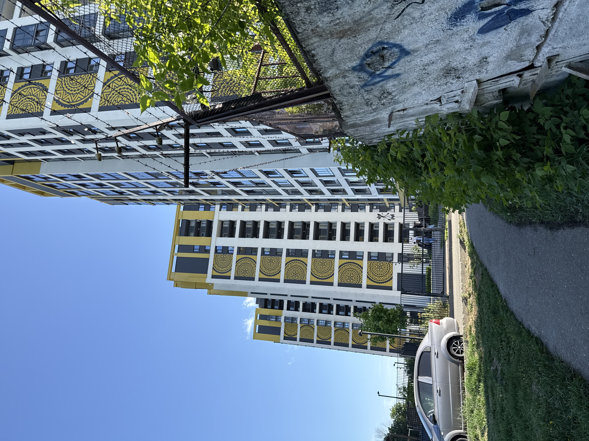 A low-angle shot captures a tall apartment building in Izhevsk, Russia, with distinctive yellow and black circular patterns on its white facade. The clear blue sky serves as a backdrop. A car is parked along a paved walkway and grassy area to the right. A wire fence with a concrete structure sits atop the building, partially obscured by foliage.