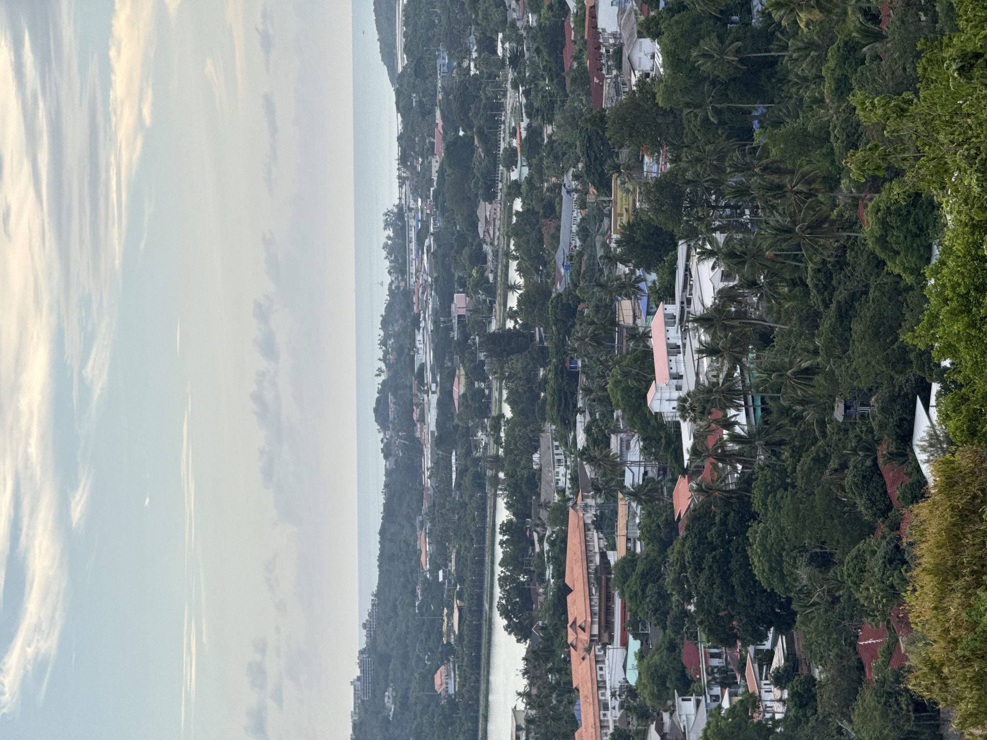 This is a high-angle, medium shot of a coastal town in Ko Samui District, Thailand. The scene is dominated by lush green vegetation and numerous buildings with red-tiled roofs, indicative of tropical architecture. In the mid-ground, a body of water, possibly a lake or lagoon, is visible, surrounded by trees and more structures. Further in the background, the ocean stretches to the horizon, meeting a pale, cloudy sky. The overall impression is of a densely populated, tropical urban landscape nestled amongst nature. There are no people or animals clearly visible in the image. The image appears to have been taken during daylight hours, possibly late afternoon or early morning, given the soft lighting.