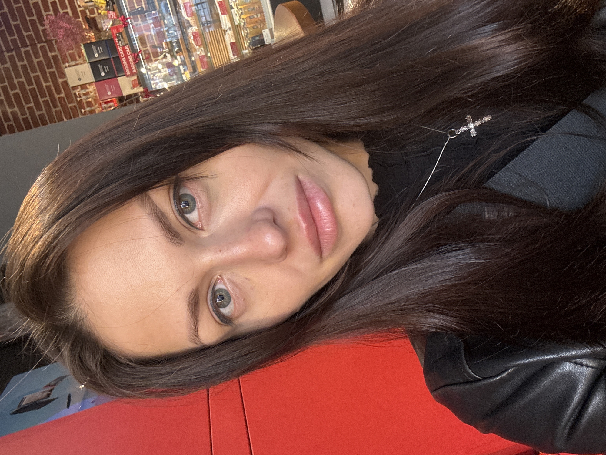 A medium close-up shot features a woman with long, dark brown hair and fair skin. She is wearing a black top and a black leather jacket. A silver cross necklace is visible around her neck. Her face is slightly tilted towards the camera, revealing her green eyes. The background shows a blurred interior, possibly a cafe or shop, with brick walls, shelves with packaged goods, and other items out of focus. The image is taken at an angle, and a red surface is seen in the lower part of the frame. There is no indication of location in Murino, Russia.