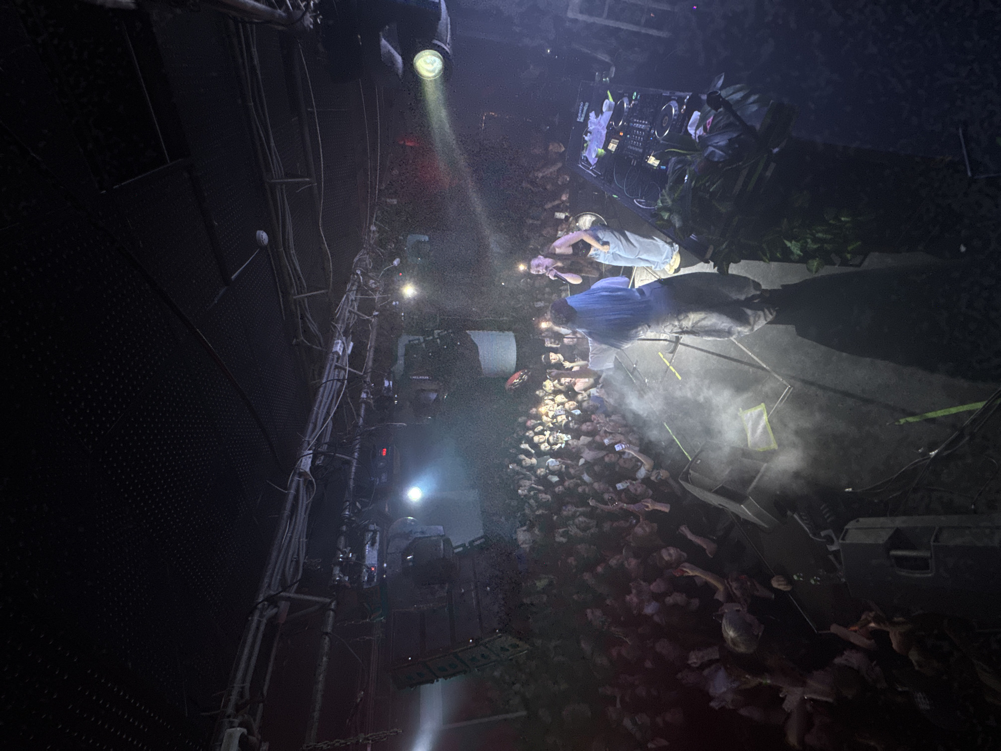 A crowded nightclub in Kazan, Russia features a stage with two performers. A man in a blue shirt is speaking into a microphone, while a woman in a similar outfit is also holding a microphone. The crowd is reaching towards the stage, illuminated by a spotlight. DJ equipment sits on a nearby platform, surrounded by foliage. The venue's interior has exposed metal structures and rigging.