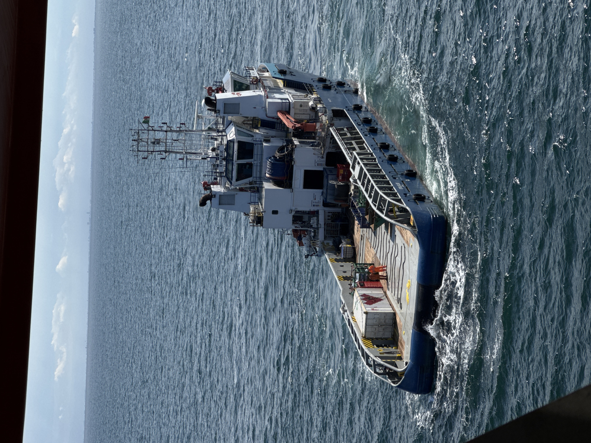 A high-angle shot depicts a large supply vessel, possibly off the coast of an Italian city, situated on the open ocean under a partly cloudy sky. The vessel, predominantly white and blue, is equipped with multiple decks, a prominent bridge, and a support structure featuring various antennas. The deck carries several containers and what appear to be personnel in orange suits. The surrounding ocean is characterized by moderate waves and textured water. The angle of the shot suggests it was taken from a higher vantage point, possibly another vessel. A dark, indistinct shape is visible at the edges, suggesting it was taken from inside an enclosure. The precise location and city in Italy remain undetermined.