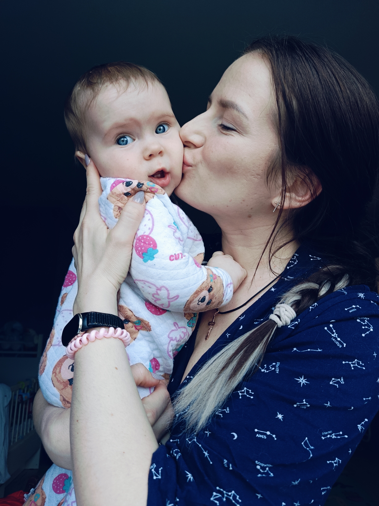 A woman is holding a baby in her arms and kissing the baby's cheek. The baby is wearing a white onesie patterned with strawberries and bears. The woman is wearing a dark blue robe patterned with constellations, and a black and pink hair tie is visible on her wrist. The background is dark and out of focus, but a white crib is partially visible. The location is unknown and cannot be inferred from the image.
