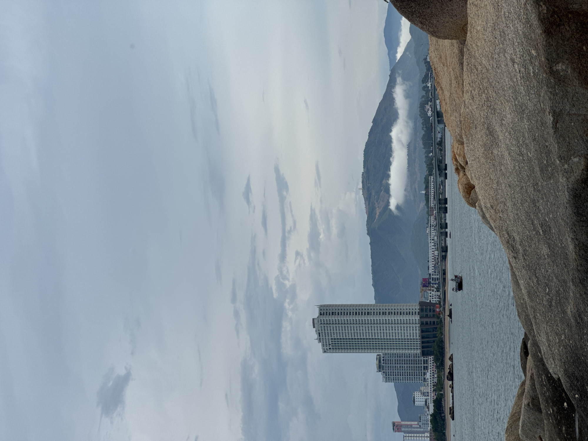 The image captures a serene coastal cityscape in Nha Trang, Vietnam, likely from a rocky vantage point overlooking the ocean. In the foreground, large, weathered rocks dominate the lower portion of the frame, their rough textures and earthy tones contrasting with the calmer waters beyond.
The middle ground reveals a bay with a small fishing boat navigating the gentle waves. The water is a muted blue-gray, reflecting the overcast sky. Along the coastline, a low-rise cityscape stretches out, featuring a series of buildings, some of which are modern high-rise apartments or hotels, characterized by their white facades and numerous windows. A prominent bridge spans across a section of the water, connecting different parts of the city, with a line of vehicles visible on its surface.
In the background, a range of lush green mountains rises majestically, their peaks partially obscured by wisps of white clouds. A prominent structure, possibly a tower or observation post, stands atop one of the distant hills. The overall atmosphere is tranquil and somewhat subdued due to the cloudy sky, evoking a sense of calm coastal living. The scene appears to be captured during daylight hours, though the overcast conditions suggest it might be early morning or late afternoon. No people are clearly discernible in the image.