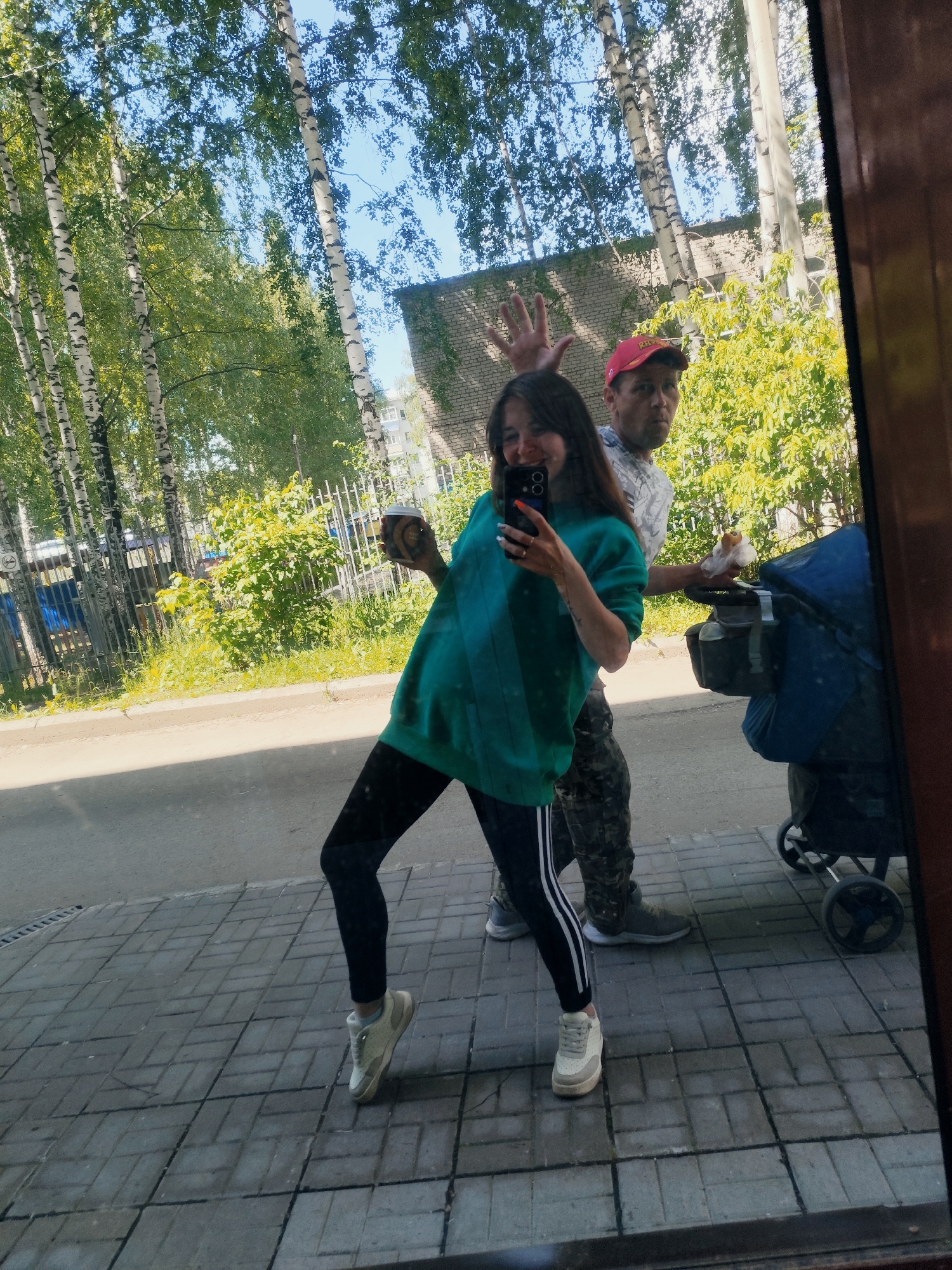 This image is a reflection, likely captured in a window or mirror, depicting two individuals outdoors on a paved surface in the city of Chaikovsky, Russia.
In the foreground, a young woman is positioned on the left, smiling at the camera. She has long dark hair and is wearing a loose-fitting teal or green long-sleeved top and black leggings with two white stripes running vertically along the outer side of her left leg. She wears light-colored sneakers. Her right hand holds a dark-colored smartphone, equipped with a dual camera setup, pointed towards herself, indicating a selfie. Her left hand holds a disposable dark brown coffee cup. She is actively posing, with her left leg slightly lifted. Her fingernails are painted, displaying both orange and a lighter shade, possibly white or light blue.
Behind and to the right of the woman stands an older man. He is wearing a red baseball cap, a light-colored short-sleeved shirt with a subtle pattern, and camouflage-patterned trousers. His footwear consists of gray athletic shoes. His right hand is raised near the woman's head, palm open, in a waving or playful gesture. His left hand is positioned near the handle of a baby stroller, holding a small item wrapped in white paper or plastic, possibly food. The stroller, visible on the far right, is blue with a dark bag or compartment attached to its handle.
The environment visible in the reflection suggests a sunny day. The ground is a grey paved sidewalk composed of rectangular blocks, bordered by a darker asphalt road. Beyond the paved area, numerous birch trees with distinctive white bark and green foliage are prominent, with bright blue sky visible through their leaves. A metal fence separates the sidewalk area from a grassy section. In the background, a light-colored brick building with windows is partially visible behind the trees and the man.