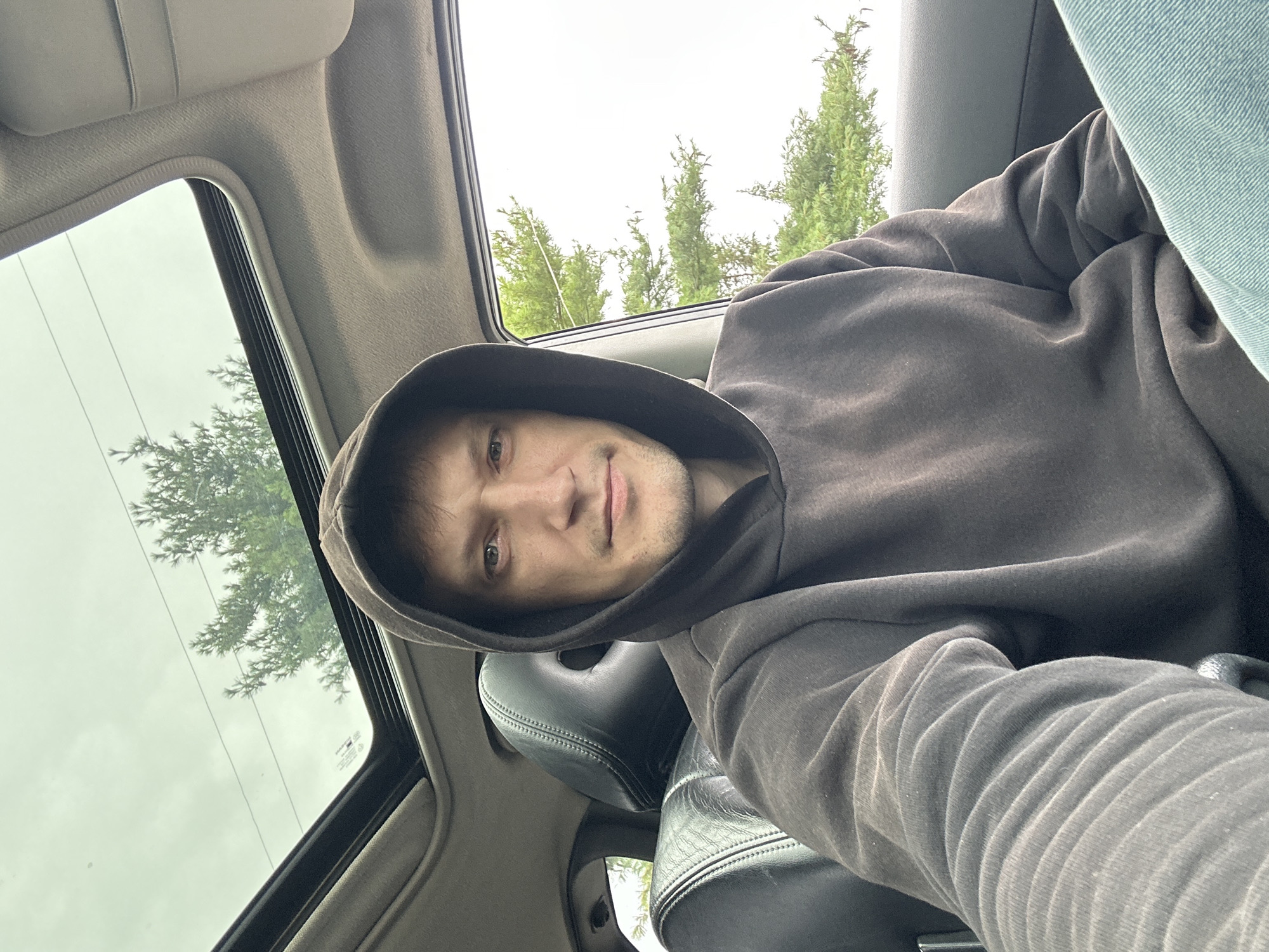 A young man with short, dark hair and light skin is seated in the driver's seat of a car. He is wearing a dark grey hooded sweatshirt and blue jeans. The hood of the sweatshirt is up, framing his face. He is looking directly at the camera with a neutral expression. The car has a sunroof, which is open, revealing a cloudy sky and the tops of green trees. The interior of the car is visible, including black leather seats. The scene appears to be in Makiv, Ukraine, given the context.
