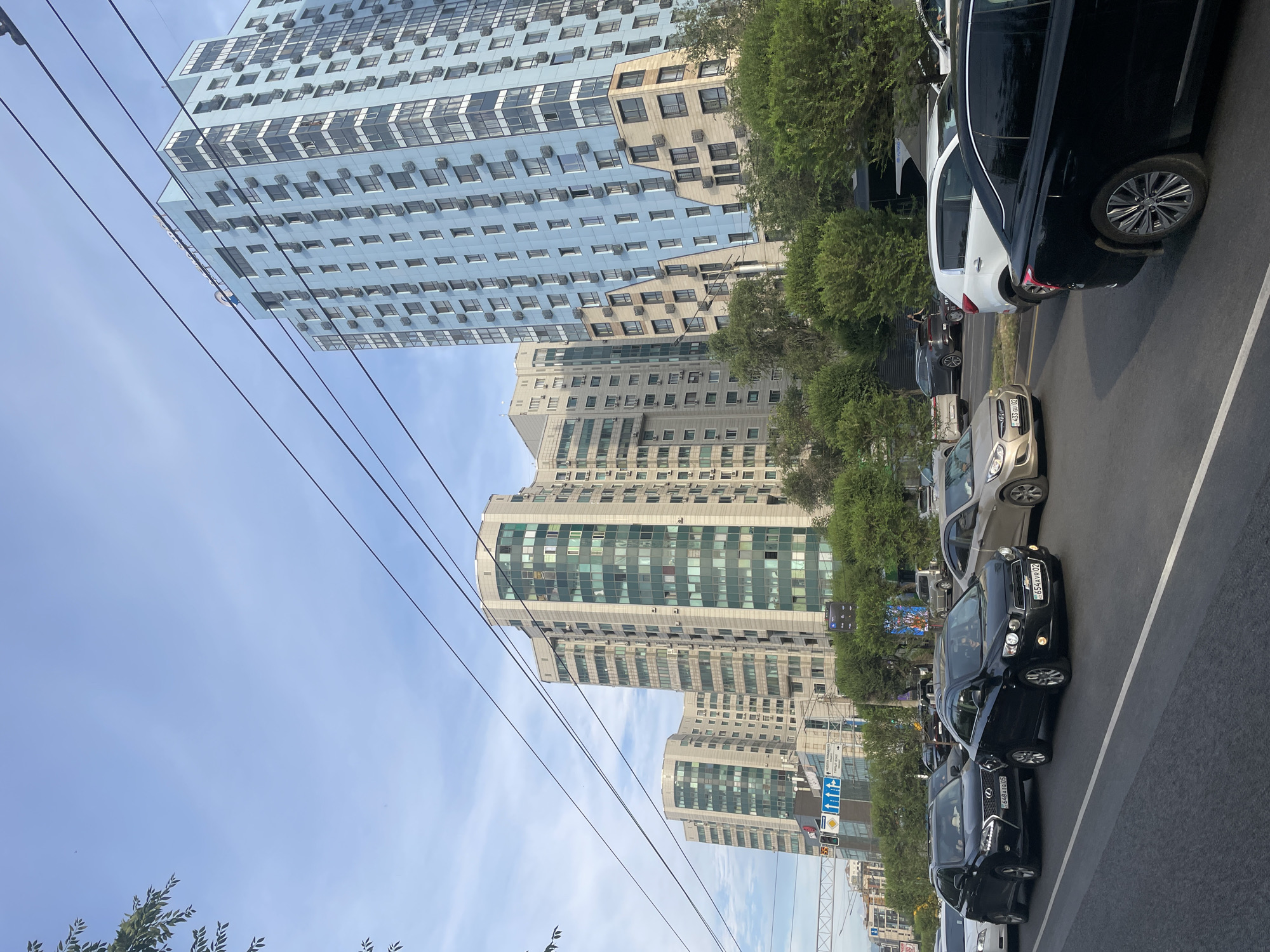 The image captures a street scene in Almaty, Kazakhstan, with modern high-rise buildings dominating the background. The buildings are characterized by their numerous windows and visible air conditioning units, suggesting a contemporary urban environment. Several cars are parked or driving along the asphalt road, which is marked with white lines. Trees and greenery are interspersed along the street, providing a contrast to the concrete structures. Overhead, power lines stretch across the frame, indicating infrastructure typical of a city. The sky is a clear blue with some wispy clouds, suggesting it is daytime, likely a sunny day. The overall impression is of a busy, well-developed city with a mix of residential and commercial buildings. No specific event or activity is clearly depicted, other than the presence of vehicles and buildings. One of the license plates visible on a black car reads "654VRP02". Another license plate on a dark car reads "649AVR02". Road signs with arrows and traffic signals are also visible. The image appears to be taken from a slightly elevated perspective, looking down the street.