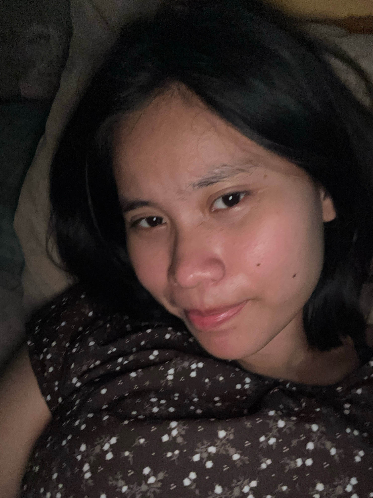 The image shows a close-up, overhead shot of a woman with dark hair lying down, likely in a bed given the presence of a pillow. She is wearing a dark brown dress with a white floral pattern. Her facial features are visible, including her eyes, nose, and mouth. Her skin appears to have a warm tone, and she has a mole on her right cheek. The lighting is dim, suggesting the photo was taken indoors in low light conditions. There is no discernible background that would indicate a specific location within East Jakarta, Indonesia.