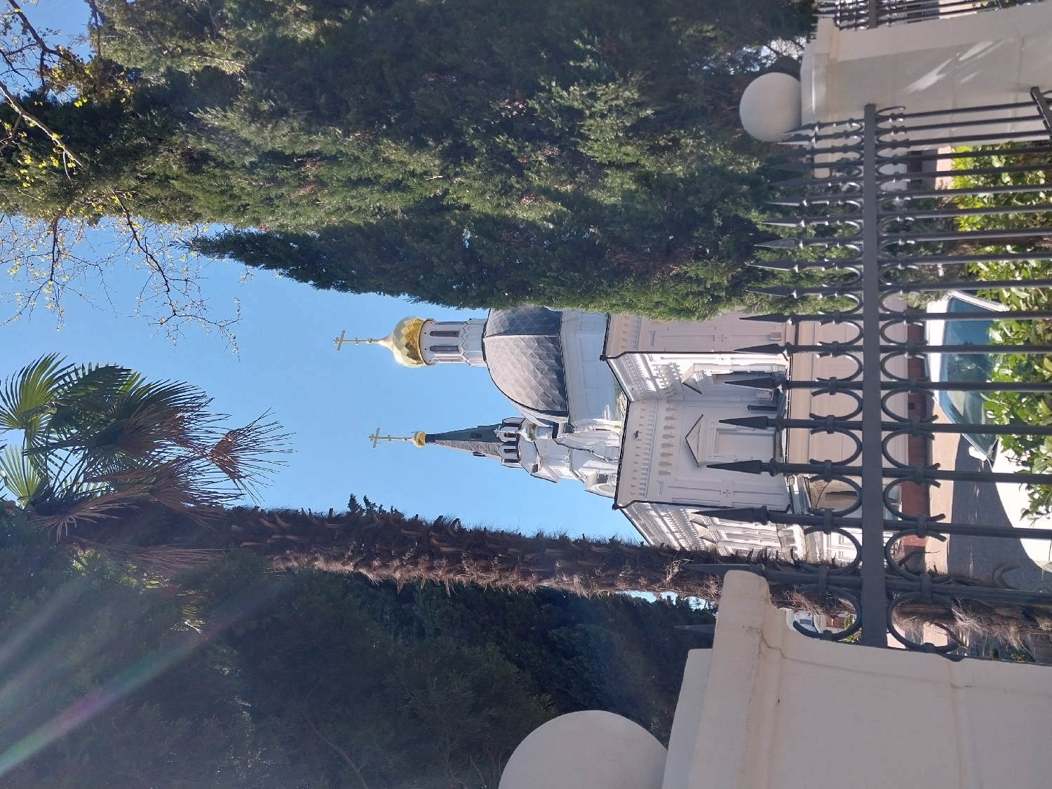 The image depicts a serene outdoor scene centered around a white Orthodox church under a clear blue sky. The church features multiple golden onion-shaped domes, each topped with a cross, and dark grey roofs. Its white facade is adorned with intricate architectural details and multiple windows.
In the foreground, an ornate dark metal fence with pointed finials and decorative circular elements partially obstructs the view of the church. To the left, a tall palm tree with a thick, textured trunk and broad green fronds stands prominently. Dense green coniferous trees, possibly cypresses, form a backdrop behind the church and line the upper part of the frame. Some bare tree branches are also visible against the sky. Portions of a white balustrade or wall, along with a spherical white decorative object, are visible in the lower right foreground.
The scene suggests a sunny day with bright, clear weather, likely midday or early afternoon, given the intensity of the blue sky and natural light. There is no human activity visible. The architectural style, particularly the golden onion domes and crosses, strongly indicates an Orthodox Christian place of worship. The presence of palm trees and coniferous trees points to a subtropical climate, consistent with the provided context of Sochi, Russia. No visible text is present in the image.