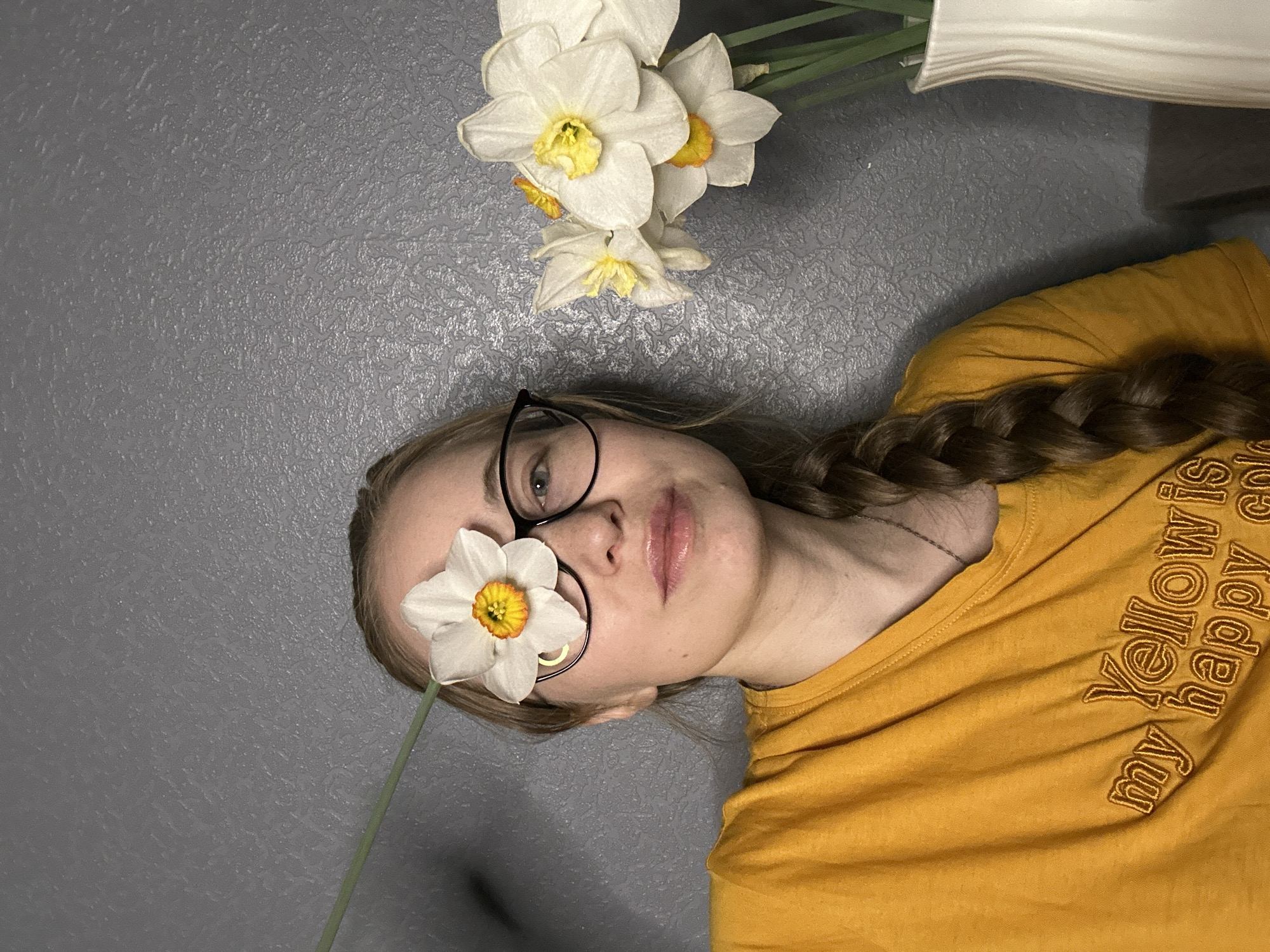 A woman with blonde hair braided over her right shoulder, is wearing a mustard yellow t-shirt with the words "my Yellow is happy color" embroidered on the sleeve. She is lying on a gray textured surface. A single white daffodil flower with a green stem covers her left eye. She has on black framed glasses. Above her head are several more white daffodils in a white vase. The location in the city of Saint Petersburg, Russia is not discernible from the image.