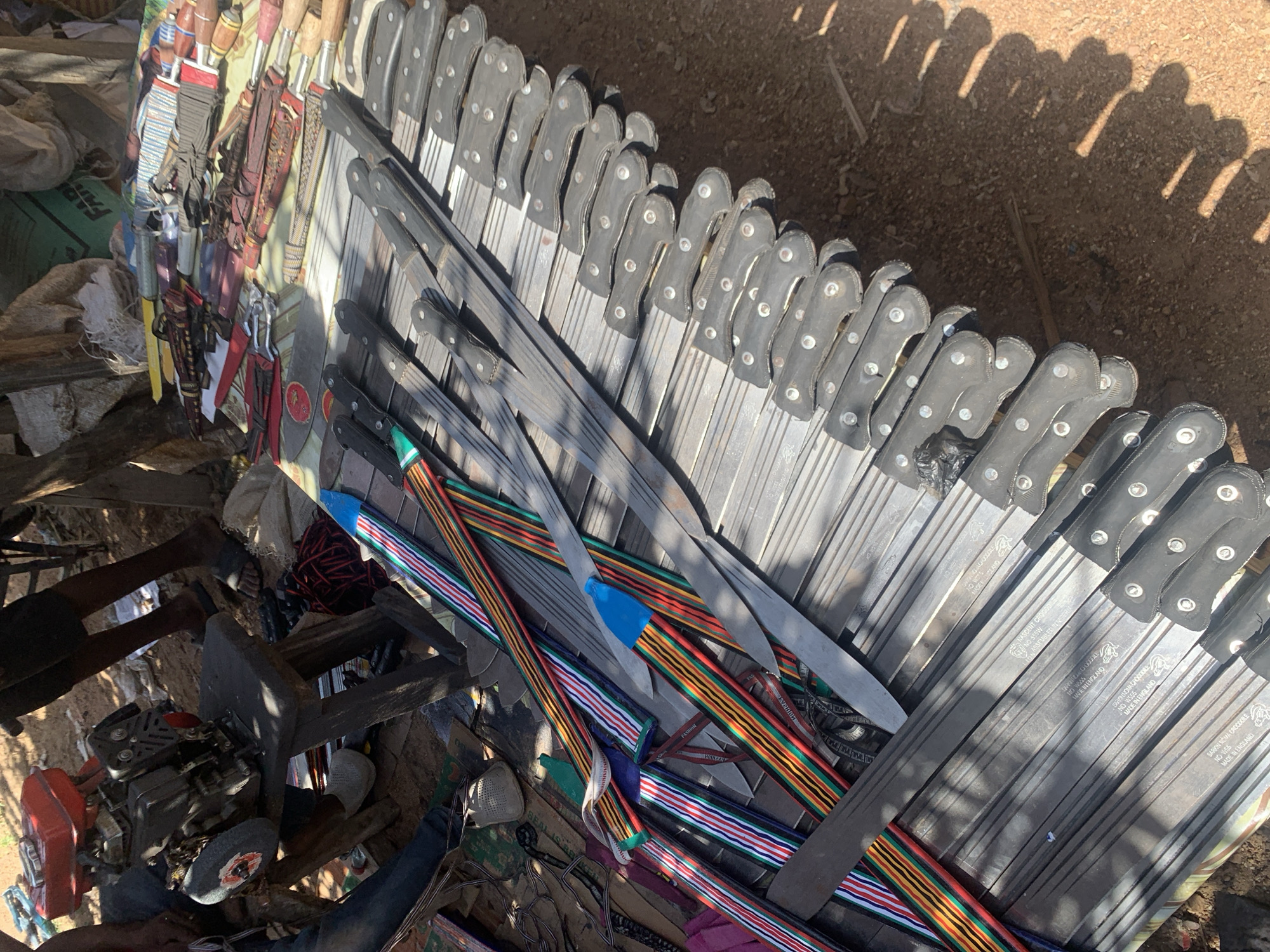 This media file shows a market stall in Nigeria, displaying a variety of items, predominantly knives and tools. The scene is set outdoors, with the ground appearing to be dirt and sand, suggesting a dry, possibly sunny environment, as indicated by the strong shadows.
The primary subjects are numerous knives, arranged in neat rows, with their blades facing towards the right side of the frame. These knives appear to be of various sizes and designs, some with black handles and others with more decorative, patterned handles featuring stripes of red, yellow, green, blue, and white. Some knives are sheathed in colorful, striped fabric. In the foreground and background, there are other objects, including what appears to be a small engine with a grinding wheel, some fabric, and bundles of string or rope. There are also hints of human presence, with legs visible at the bottom left of the frame, suggesting people are present at the market.
The activity taking place is likely the sale of these items at a market. The sheer number of knives suggests they are being offered for sale, possibly for agricultural use or as tools. The time of day appears to be daytime, given the strong sunlight and distinct shadows cast by the objects, indicating a clear day. The overall impression is one of a bustling outdoor market where tools and practical items are being traded.
Visible text on the knives includes "MADE IN ENGLAND," "NO 555W," "NASCENT CROOD," and "SARKON NOMA CROCODILE," suggesting the origin and possibly branding of some of the tools. The presence of "Project, Nigeria" in the location context further solidifies the setting as a market in Nigeria.