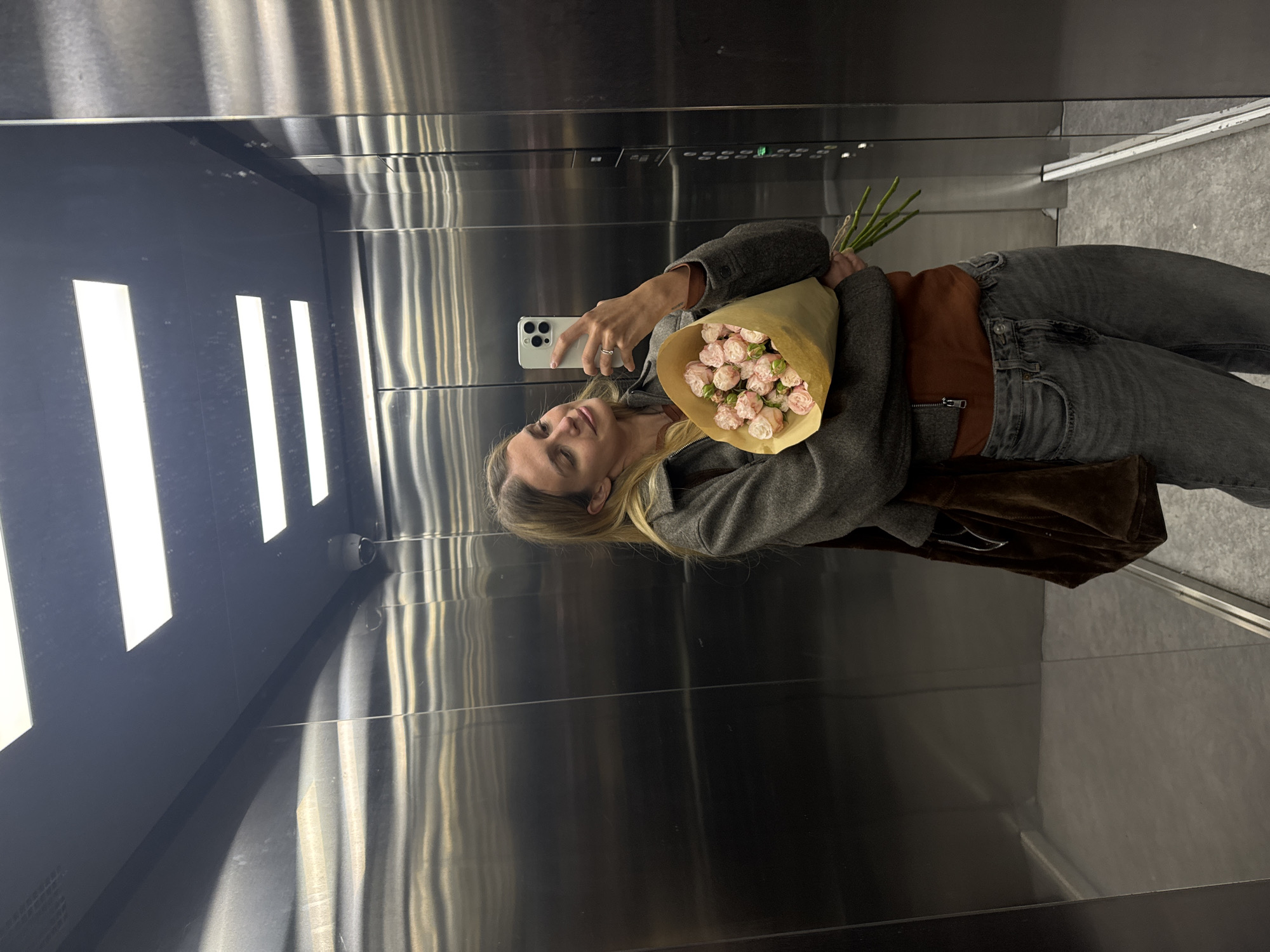 A woman stands in an elevator holding a bouquet of pink roses. She is taking a selfie with a smartphone. The elevator has stainless steel walls with vertical ridges. A strip of LED lights runs down one side of the elevator. The floor is gray. The location is Unknown, Unknown.