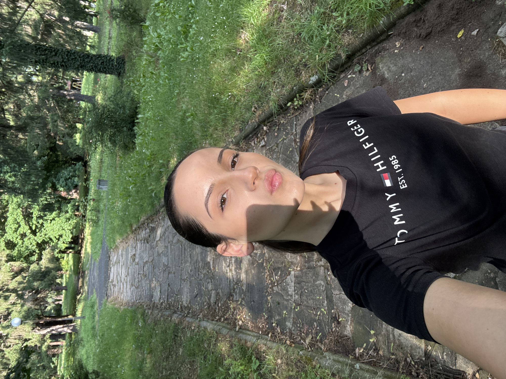 The image displays a close-up selfie of a young woman taken outdoors, with the photograph appearing rotated 90 degrees counter-clockwise. The woman's head is tilted, and she is looking upwards towards the camera with a slight pout or 'duck face' expression. She has dark hair pulled back, brown eyes, and a healthy complexion, with a distinct shadow cast across the left side of her face, indicating strong sunlight. Another shadow is visible on her right shoulder.
She is wearing a black, short-sleeved t-shirt. Clearly visible white text runs vertically down the front of her shirt, reading "TOMMY HILFIGER," followed by a small rectangular logo (red, white, and blue segments), and then "EST. 1985."
The background reveals a natural setting, likely a park or green space, under bright, sunny weather. A rustic stone path, composed of irregular paving stones, runs diagonally across the frame from the bottom right to the middle left. The path is flanked by lush green grass, various types of trees, and dense foliage, creating a serene environment. Some areas of exposed soil and scattered small stones are visible alongside the path.