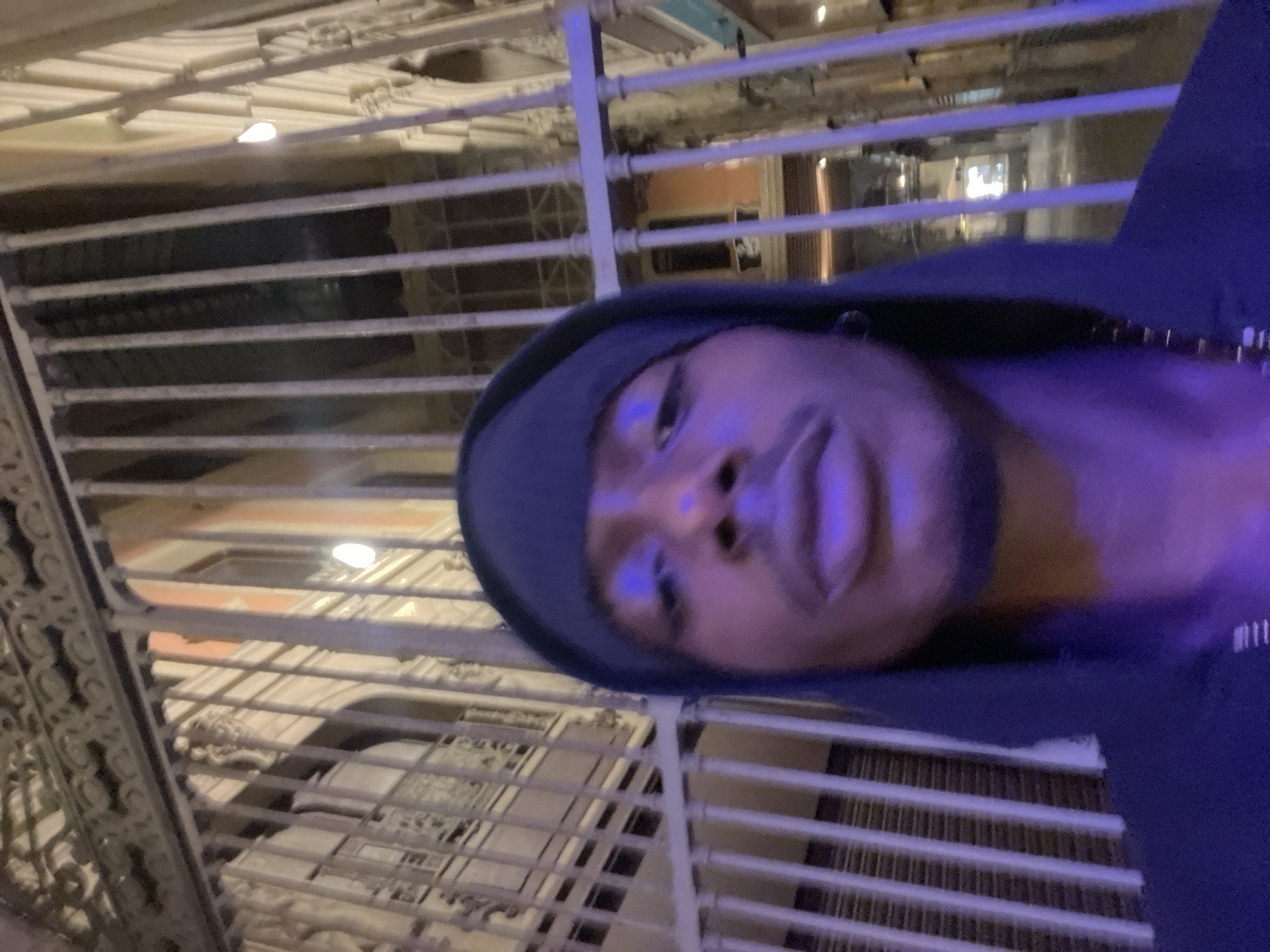 The image is a vertical selfie taken by a person of color, facing the camera with a casual expression. The individual is wearing a dark blue or black hoodie with the hood up, possibly covering a beanie, and is seen from the chest up. Their face is illuminated by a prominent purplish light source, highlighting their features.
The setting appears to be an indoor or covered architectural space, likely at night or in the evening, indicated by the artificial lighting. Behind the person, a distinctive, elaborate white or light-colored decorative railing or grate structure dominates the background. This ornate structure, possibly made of wrought or cast iron, features intricate curvilinear and geometric patterns, suggesting a significant architectural style. Multiple horizontal bars are visible, forming a grid-like pattern that could be part of a balcony, staircase, or a decorative facade of an interior courtyard or atrium. Through these bars, other building details, windows, and additional light sources are faintly visible in the deeper background.
The architectural elements, particularly the detailed, decorative ironwork, hint at a historic European building. Given the provided context of Barcelona, Spain, these features are highly suggestive of the Modernista architectural style, prevalent in the city. The person is actively taking a selfie in what appears to be a grand and ornately designed interior. There is no visible text in the image.