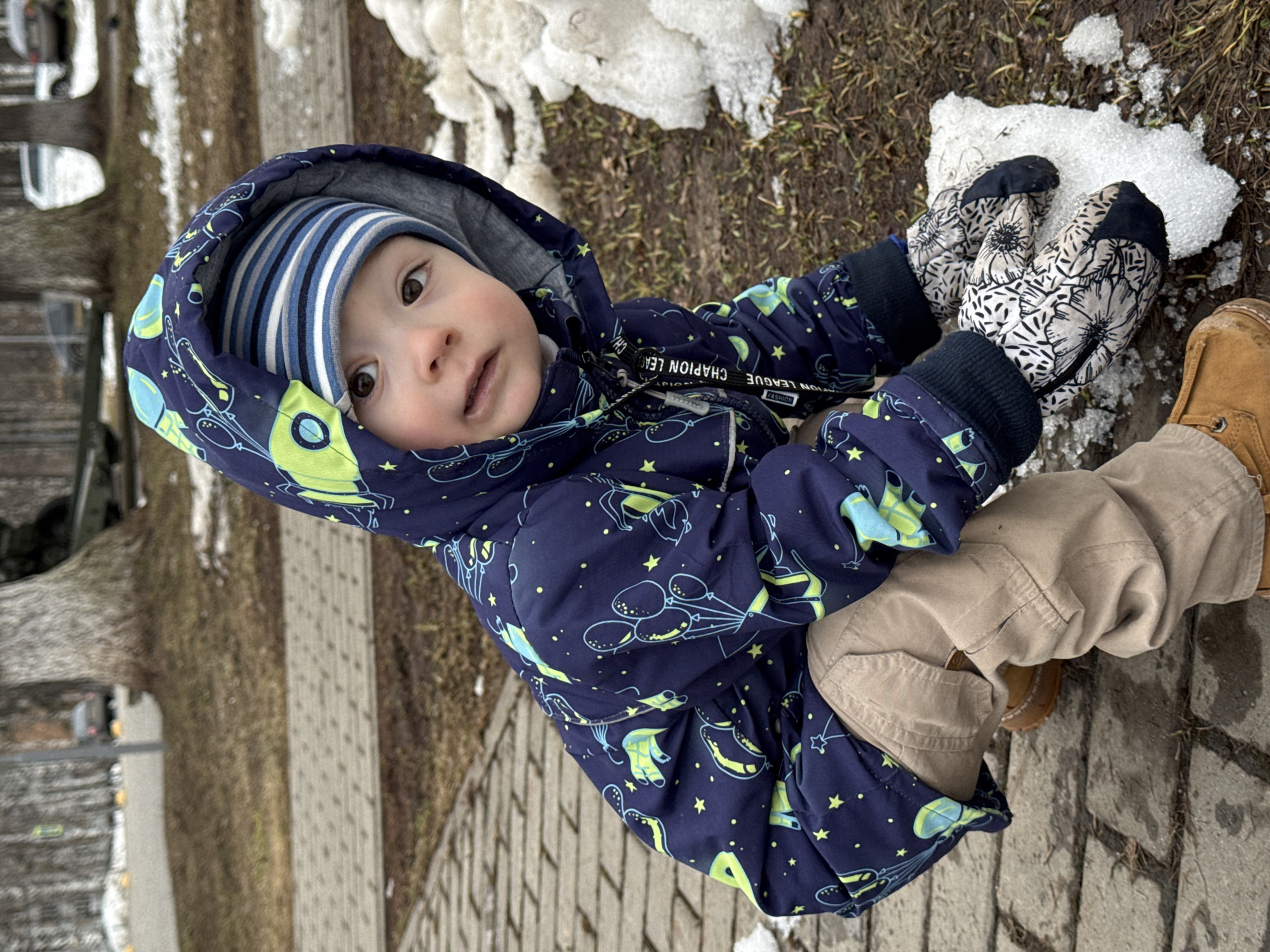 A young child in a blue hooded jacket and striped hat plays with snow. The child wears mittens and tan pants and sits on a paved surface near a patch of grass and snow. The photo was taken in the city of Izhevsk, Russia.