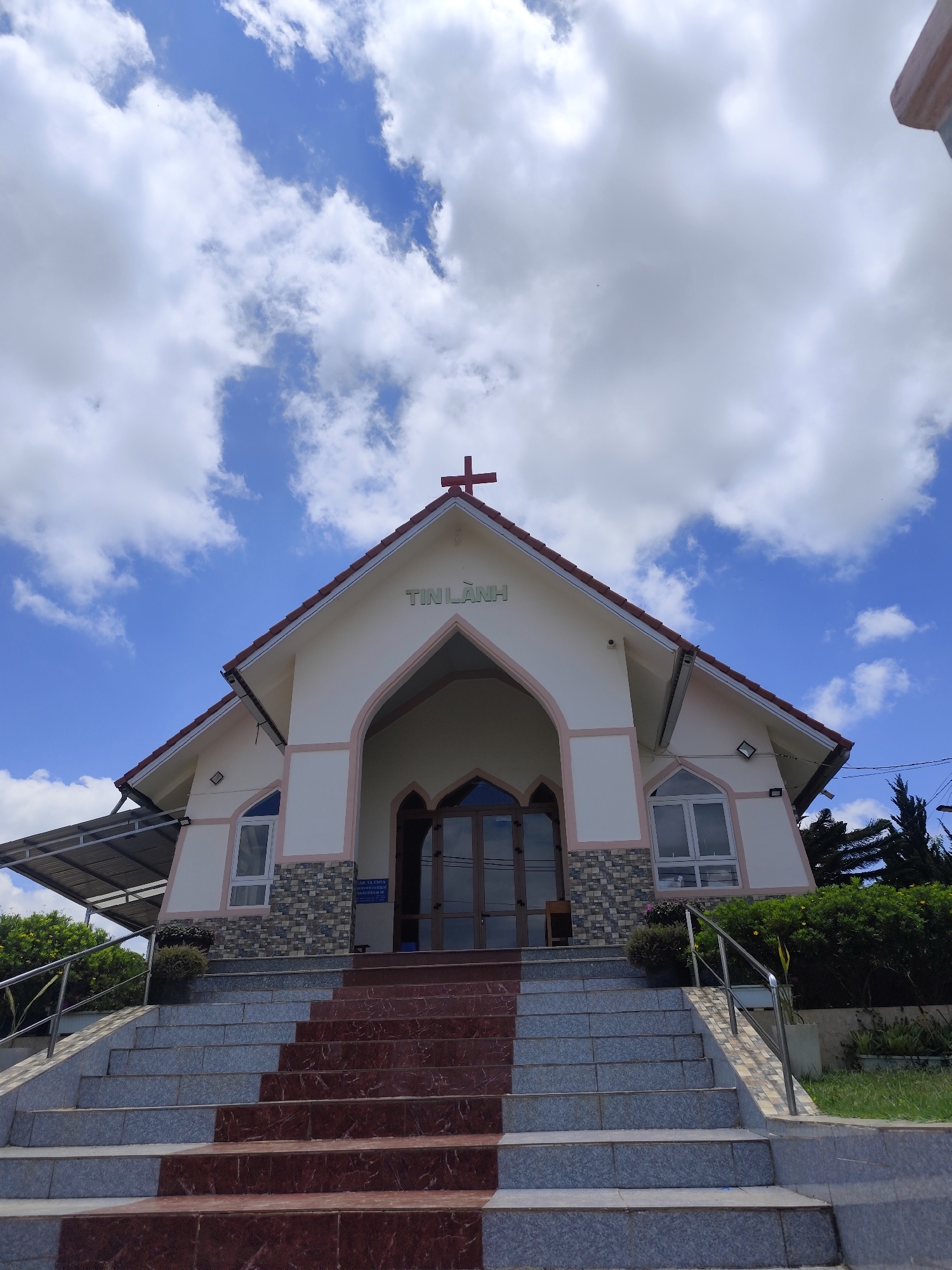 The image shows the front exterior of a church in Lạc Dương, Vietnam, on a bright day with a blue sky and scattered white clouds. The church has a pointed roof with a red cross at the peak, and the words "TIN LÀNH" are printed on the building beneath the cross. The church's facade is light-colored with pink trim around the arched entrance and windows. The lower portion of the church's walls features a stone texture. A series of stone steps, partially colored with red tiles, leads up to the church's entrance, and there are plants and railings on either side of the steps. An awning extends from the left side of the church building.