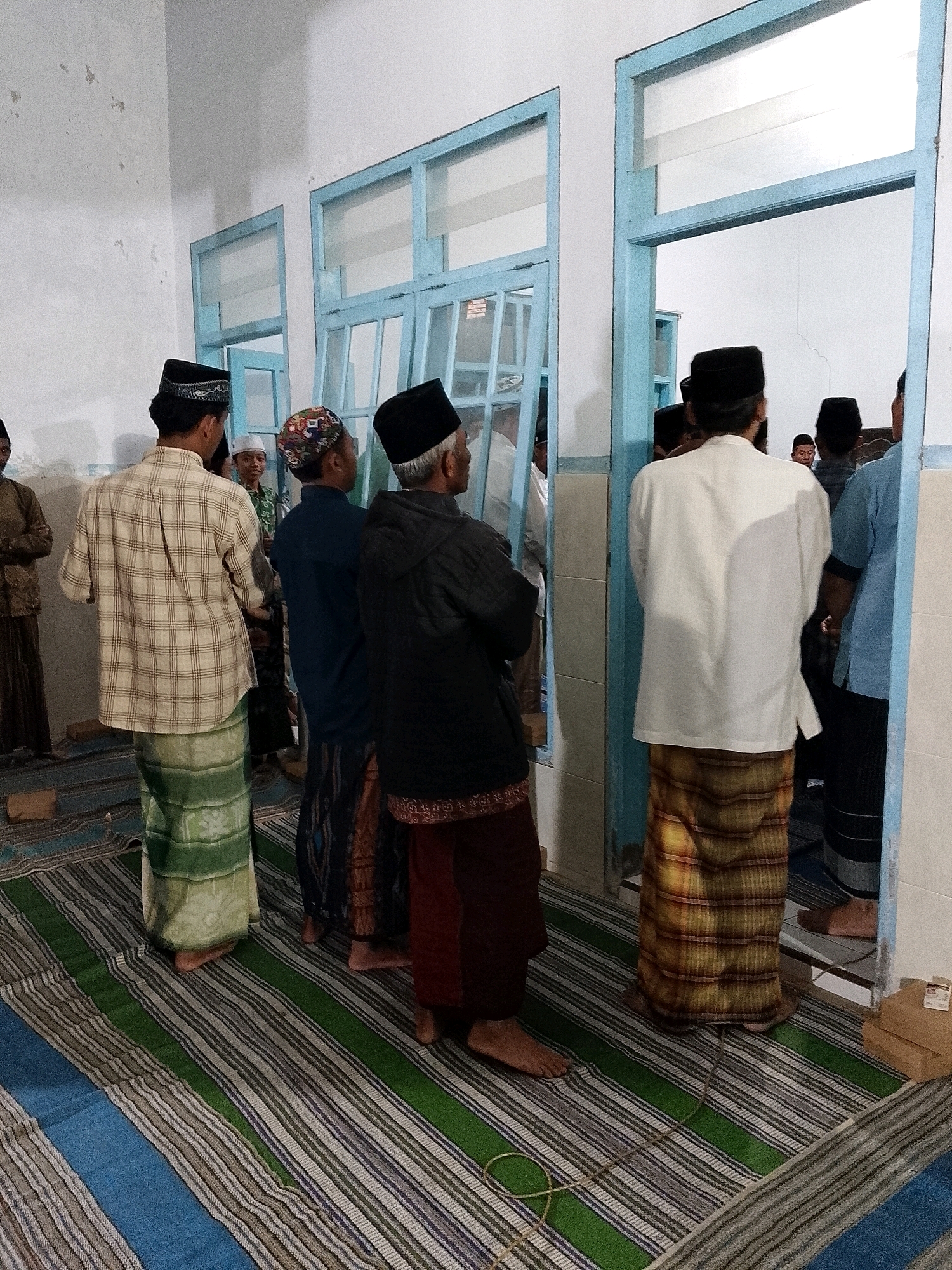 In Karangsari, Indonesia, a group of men, predominantly wearing traditional Muslim attire including sarongs and peci caps, are gathered indoors. The scene appears to be within a room with a light-colored wall and blue-framed windows or doorways. The men are standing on a striped mat that covers the floor. Some individuals are facing away from the camera, while others are partially visible from the side or are in the background. The lighting suggests an indoor environment, possibly a community hall or place of worship. The overall atmosphere is one of assembly or gathering.