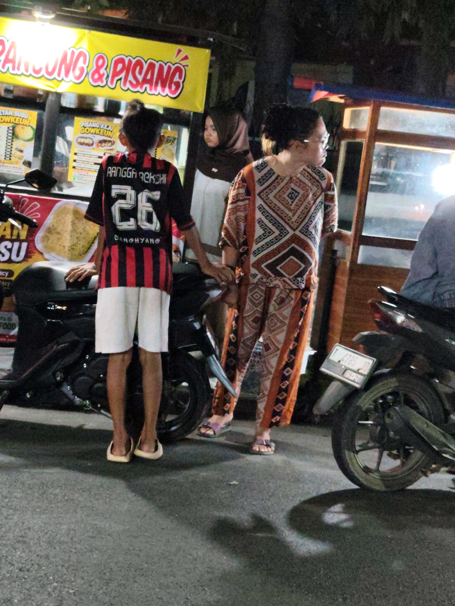 At night, in the city of Purwakarta, Indonesia, a boy wearing a red and black striped jersey with the number 26 and white shorts stands in front of a motorcycle. His jersey has the name "RANGGA REKSON" printed on the back. To his right, a woman in a patterned dress stands next to another motorcycle. Behind them, a food stall displays a yellow banner with red lettering that reads "PANGGUNG & PISANG". The stall also has various food items and menus displayed. The ground is asphalt, and shadows are cast by the lights and objects.