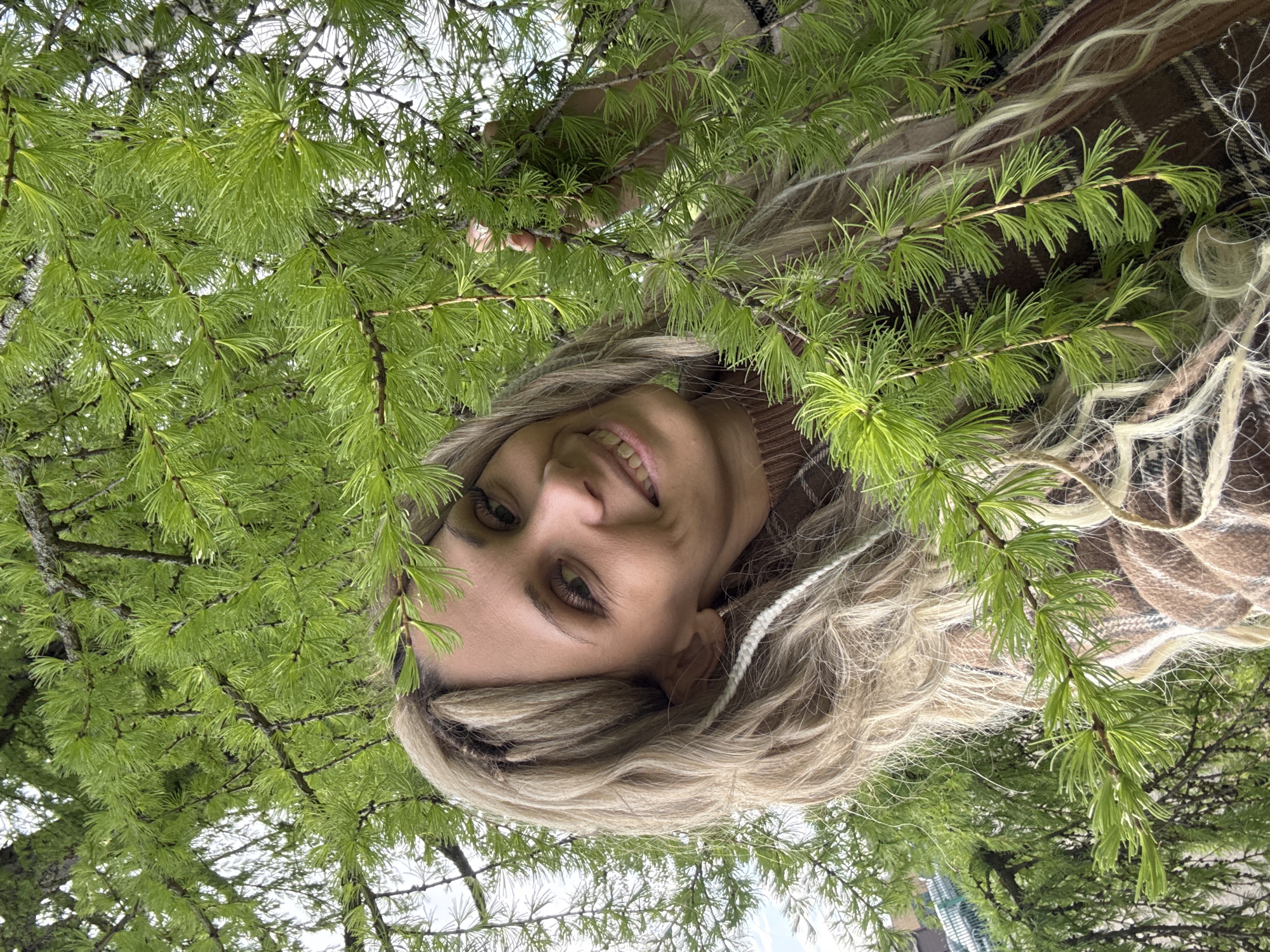 A woman with blonde hair, wearing a brown patterned garment, is seen smiling and looking directly at the camera. She is positioned amidst the branches of a green, needle-leaved tree. The photo appears to have been taken from a low angle, looking up at the woman through the tree branches. Part of a building can be seen in the background. It is possible that this image was taken in Rusanovo, Russia, but this cannot be confirmed from the image alone.
