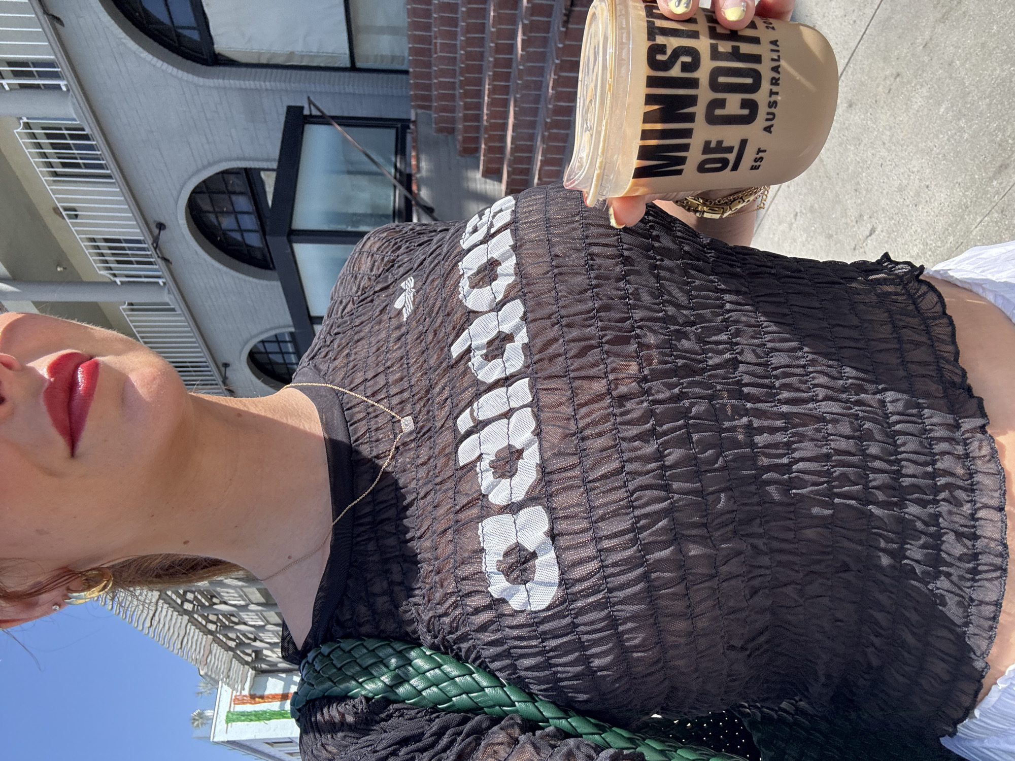 A woman stands outdoors in Santa Monica, California, holding a cup of coffee from "MINISTRY OF COFFEE". She wears a sheer black ruched top with an Adidas logo, a gold necklace with a diamond pendant, and large gold hoop earrings. Her nails are painted with a yellow and white design. A green woven bag is visible on her left shoulder. In the background, a building with arched windows and a white railing is partially visible. The ground appears to be a sidewalk and stairs.