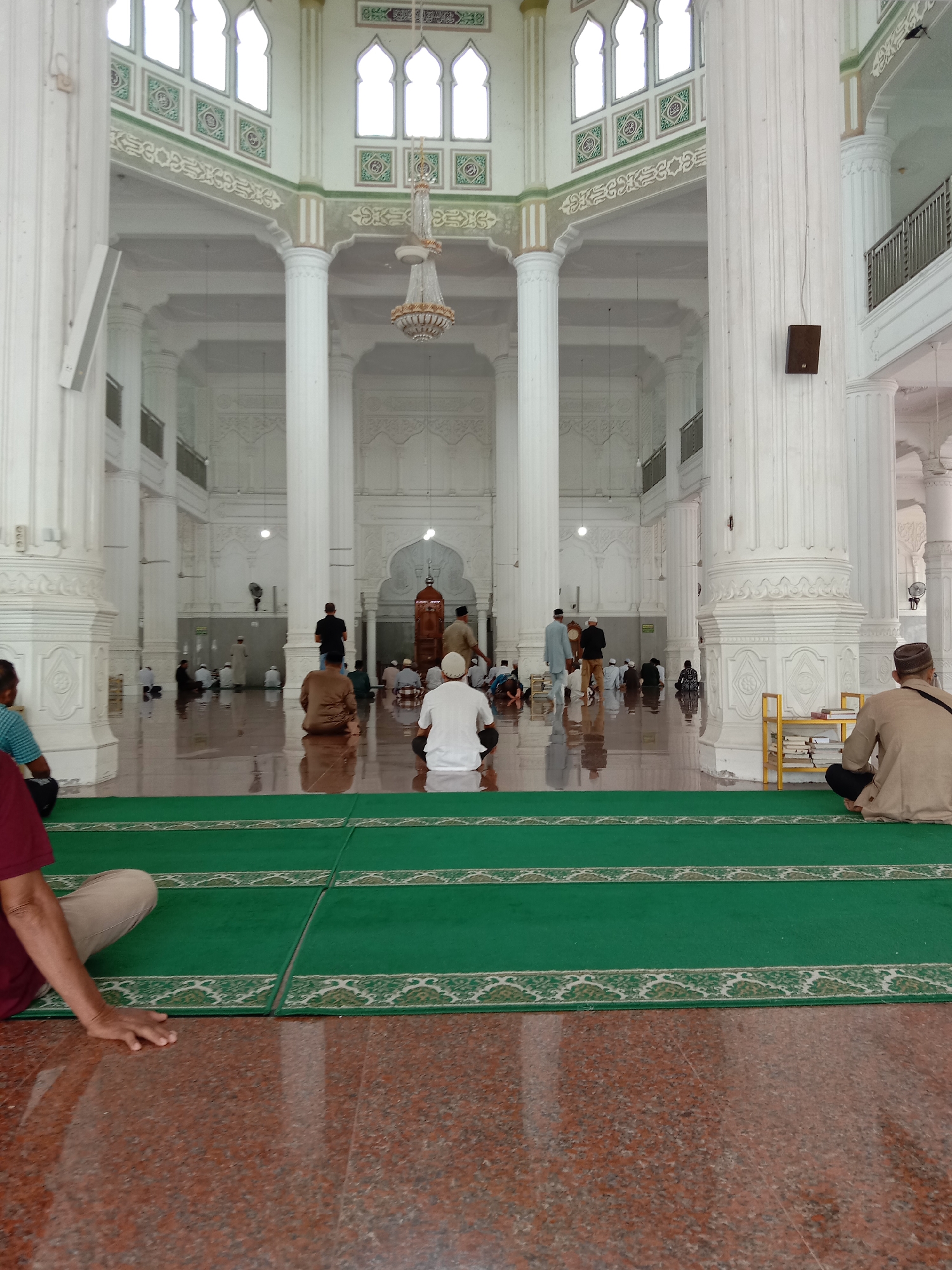 The image shows the interior of a mosque in Lhokseumawe, Indonesia. The mosque features high white columns and decorative arches, with large windows at the top. Several people are seated or standing on the reflective floor, which is partially covered by green prayer mats. The overall atmosphere is serene and contemplative.