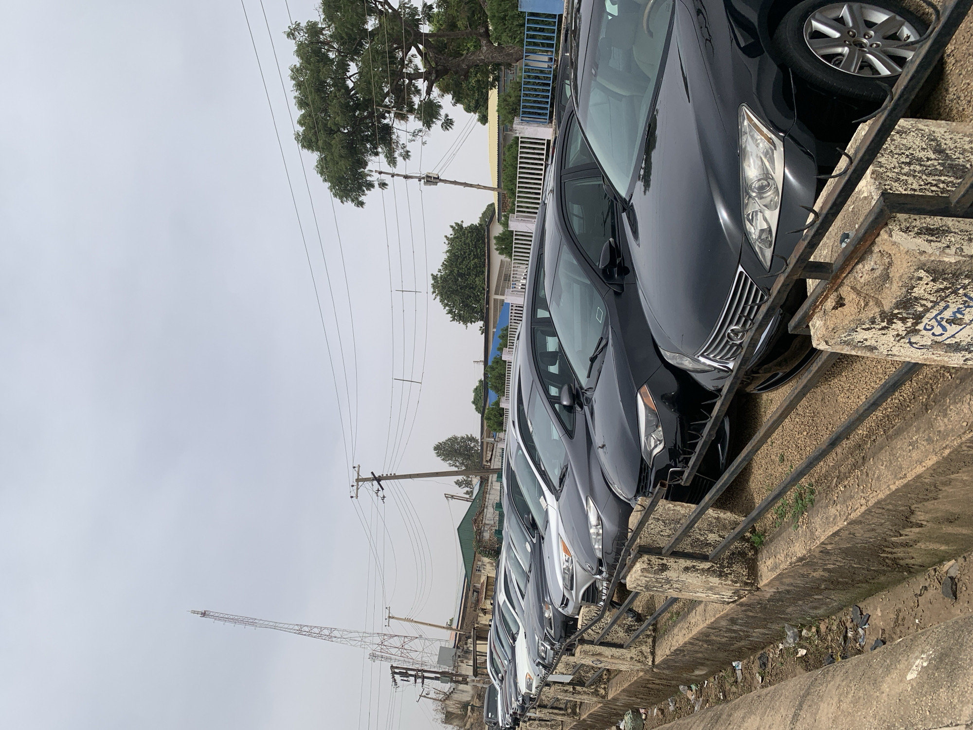 The scene shows a row of parked cars, varying in color from white to black, lined up along a concrete barrier with a black metal railing. The location appears to be an outdoor car lot or parking area. In the background, there are buildings of varied heights, power lines, a tall metal tower possibly for telecommunications, and a large tree with full foliage. The sky is overcast. The image is taken in Jos, Nigeria.