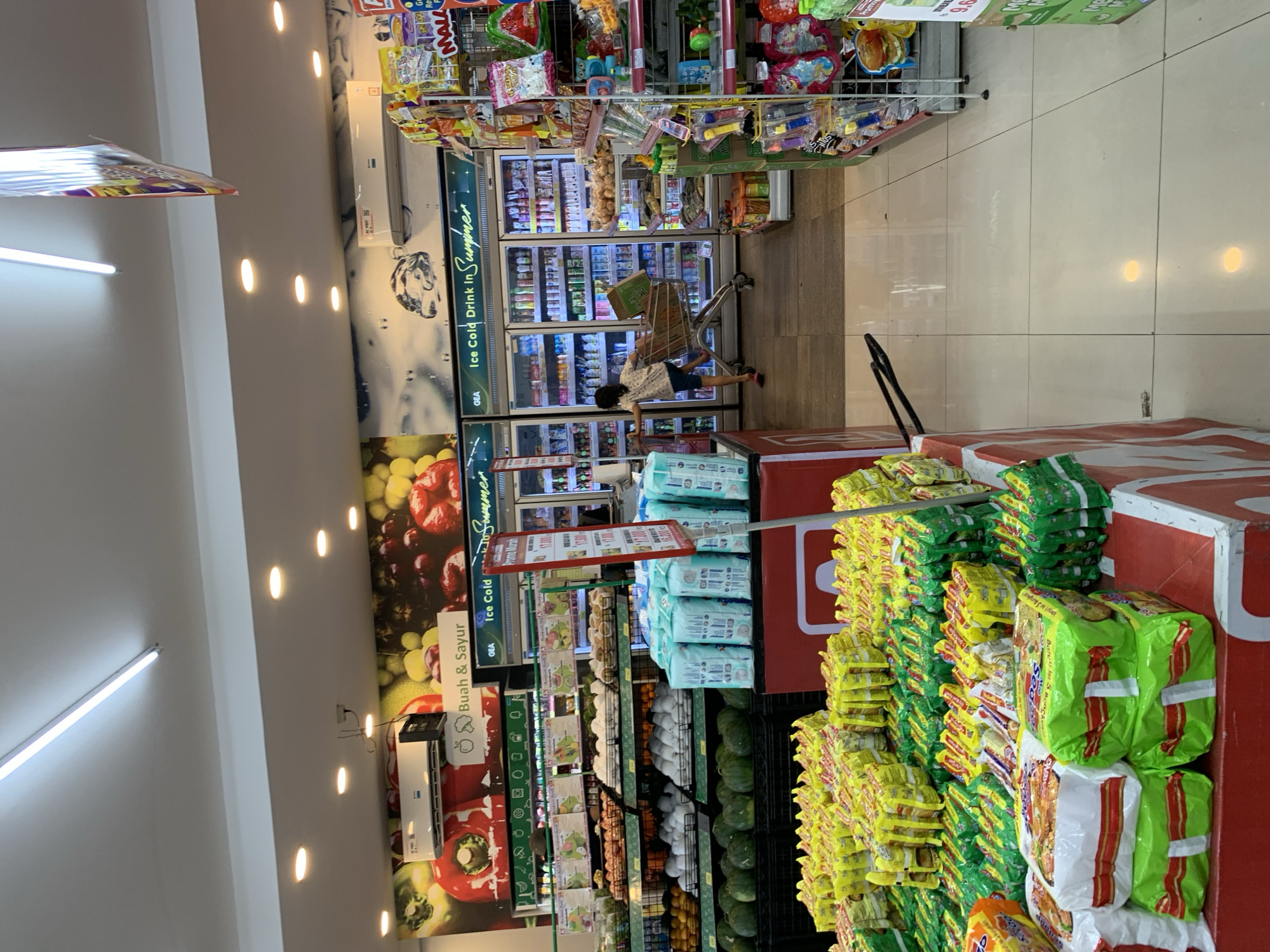 This is a vertical, eye-level shot of the interior of a grocery store or convenience store in Makassar, Indonesia. The main subjects are shelves stocked with various packaged goods, including instant noodles, bottled drinks, and produce. A young person is visible in the middle ground, pushing a shopping cart. The store appears well-lit, suggesting it is open for business, likely during daylight hours.
Dominant colors include the bright yellow and green packaging of the instant noodles that fill much of the right side of the frame, the red and white branding on a display in the foreground, and the cool blues and greens of the refrigerated drink section in the background. The shelves are neatly organized, displaying a wide variety of products. Some visible text includes "Buah & Sayur" (Fruits & Vegetables), "Ice Cold Drink in Summer," and various brand names and prices that are mostly illegible due to the image quality and angle. The atmosphere seems to be that of a typical retail environment, with no strong indicators of specific emotions or weather conditions beyond the indoor setting. The overall impression is one of a busy, well-stocked store.