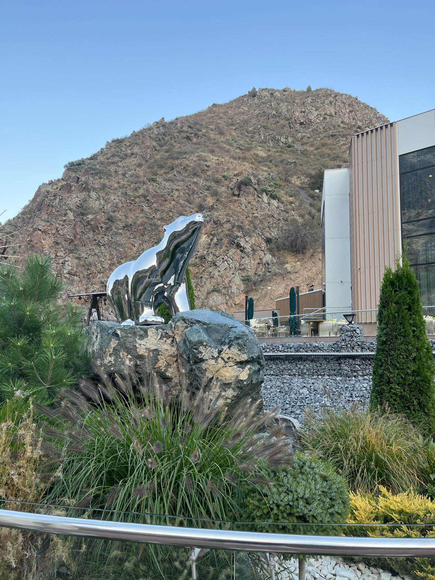 A large, polished silver bear sculpture stands on a rock formation in the foreground. Behind the sculpture, a modern building with a ribbed facade and large glass windows is visible. Further back, a dry, rocky mountain slope dominates the background under a clear blue sky. The area in front of the building features outdoor seating with tables and umbrellas. Various plants, including tall grasses and evergreen trees, are scattered throughout the scene. A metal railing runs along the bottom edge of the frame. The location is identified as Arashan, Kyrgyzstan.
