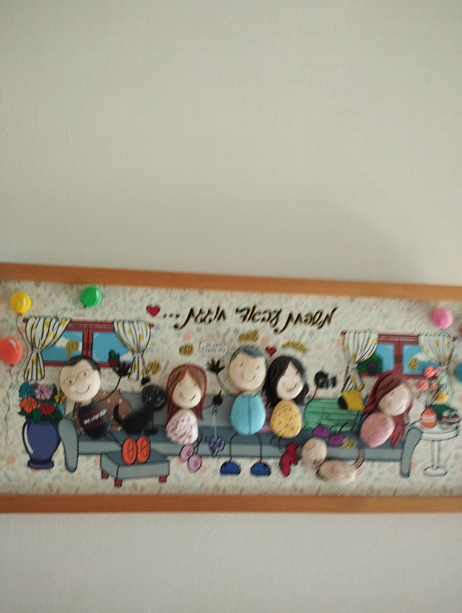 A framed artwork depicts a stylized family of five figures and a dog seated on a couch against a backdrop of a patterned wallpaper and a window. The figures, made of rounded objects, have simple facial features with dots for eyes and curved lines for smiles. The text above the figures reads "... Noah" and "New Year", written in Hebrew. The artwork is displayed on a wall in a home, likely in Pardes Hana-Karkur, Israel, suggested by the script. The overall scene is cheerful and family-oriented.