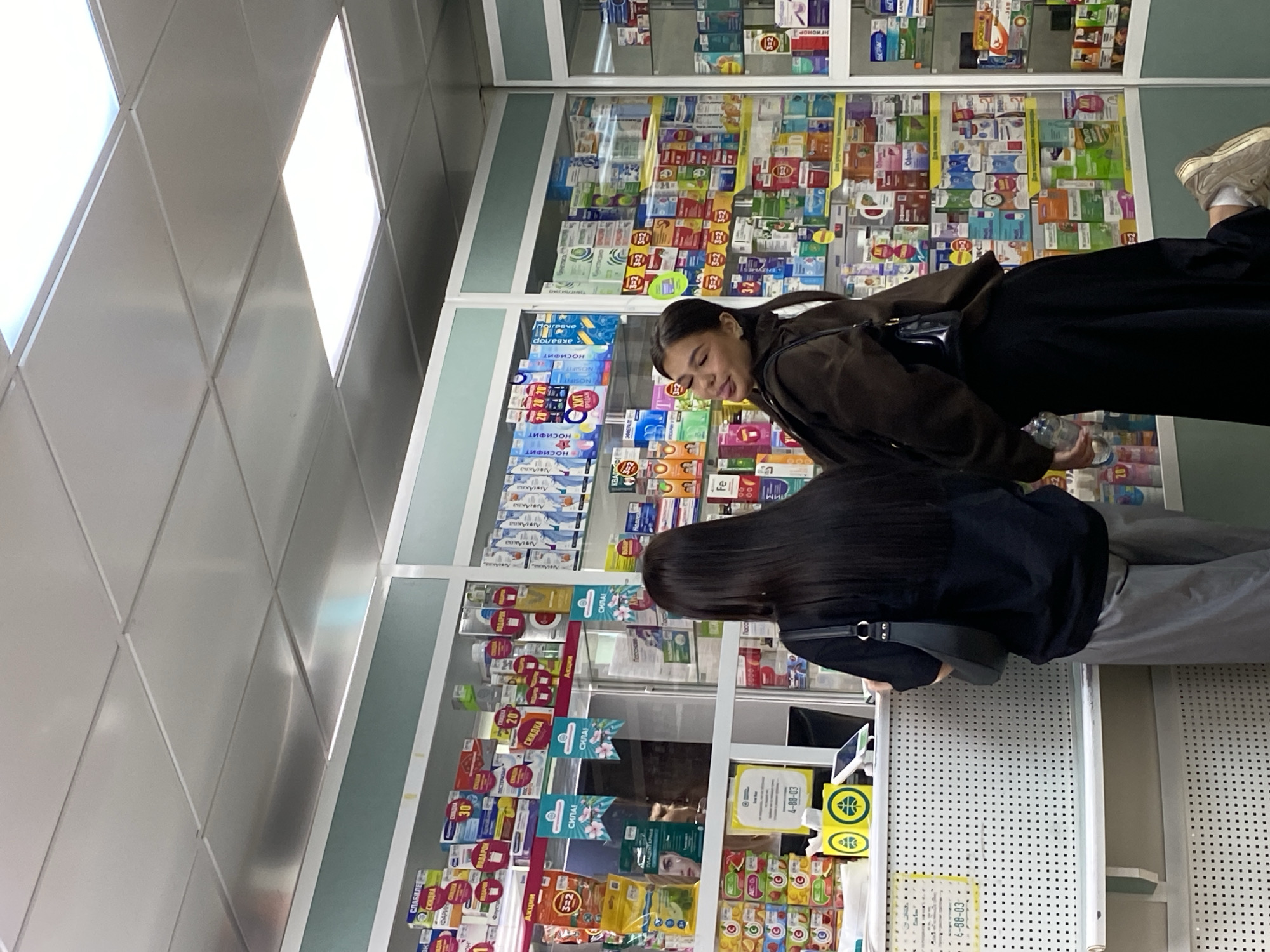 The image shows two women in a pharmacy, likely in Chaikovsky, Russia, based on the Cyrillic text on the shelves. The woman in the foreground is wearing a dark shirt and gray pants, with a black purse slung over her shoulder. She is standing at the counter. The second woman, positioned behind her, is wearing a brown jacket and black pants. She is holding a bottle of water. Shelves of medicine and other health-related products fill the wall behind them. The ceiling is comprised of diamond-shaped tiles. The pharmacy counter has signs and displays for products, including Cyrillic text indicating discounts and promotions.