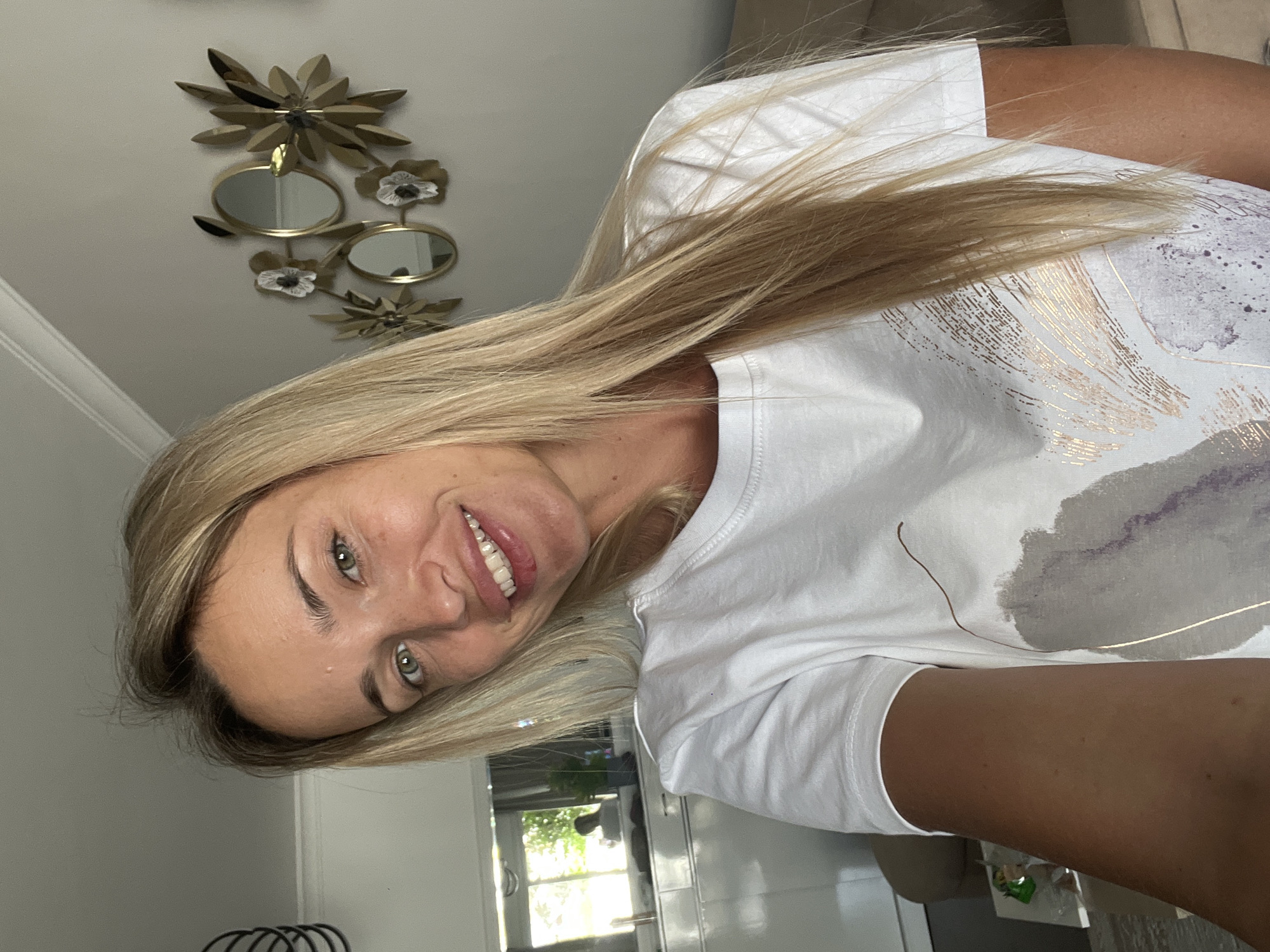 An outdoor selfie shows a fair-skinned, blonde woman in her late 20s to early 30s with green eyes and a warm smile. She is wearing a white t-shirt with a painted abstract design and her long, blonde hair falls over her shoulders. She is positioned in the foreground, and in the background, the scene appears to be indoors, possibly a living room or kitchen area. There's a reflection in a mirror showing a window and greenery outside. The woman is smiling directly at the camera. The setting appears to be a modern residential interior. There is no indication of the location being Sharm El Sheikh, Egypt.