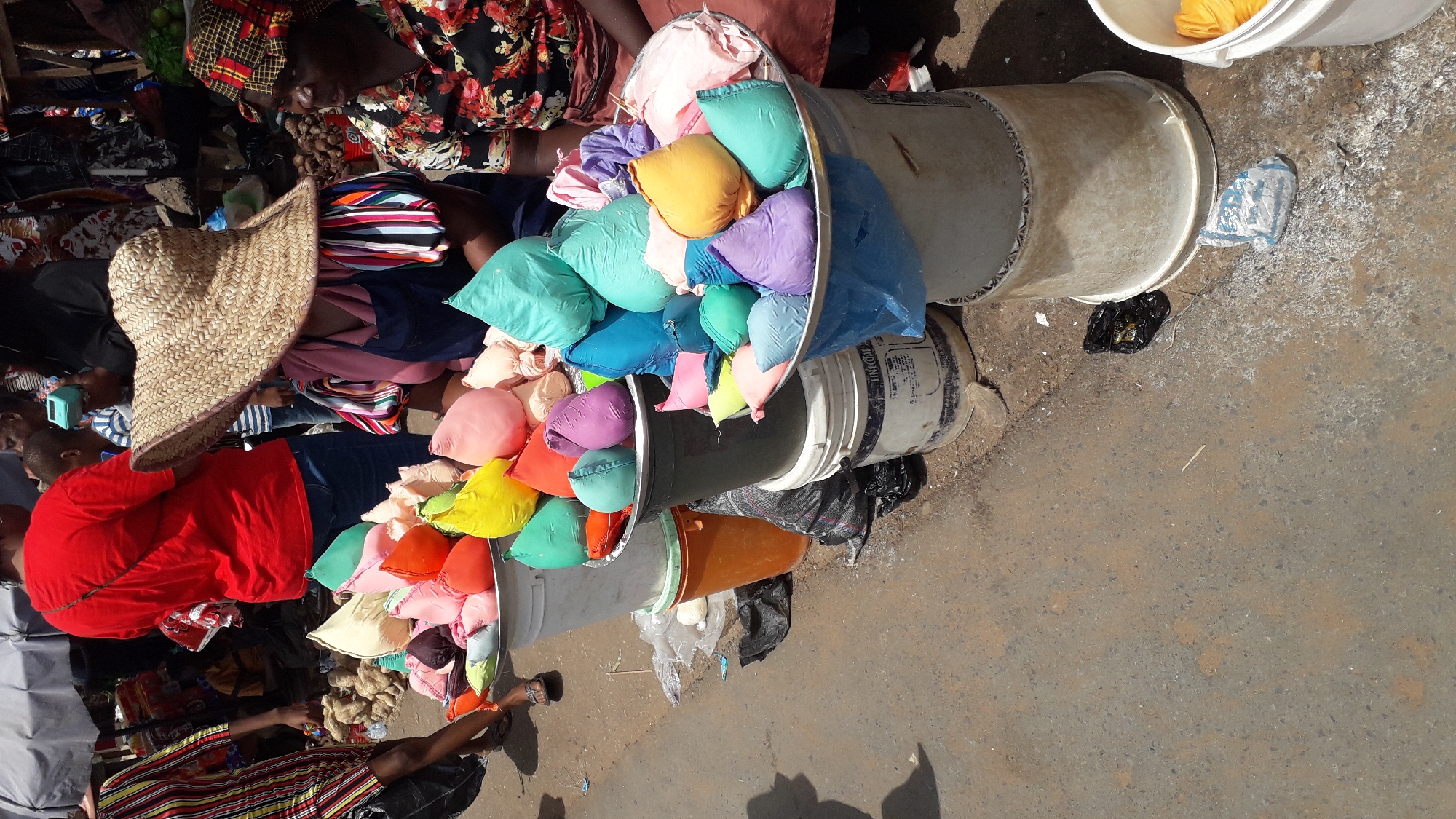 The image shows a street scene at a market in Onitsha, Nigeria. Several people are present, some seated and some partially visible. There are woven straw hats, various colored fabrics, and clothing visible. The focus is on several buckets stacked on top of each other and overflowing with small, brightly colored, pillow-like objects. These buckets are likely used for transporting or displaying these items for sale. The ground appears to be paved and somewhat dusty. Black plastic bags are scattered near the buckets. The sunlight is strong, casting shadows in parts of the scene.