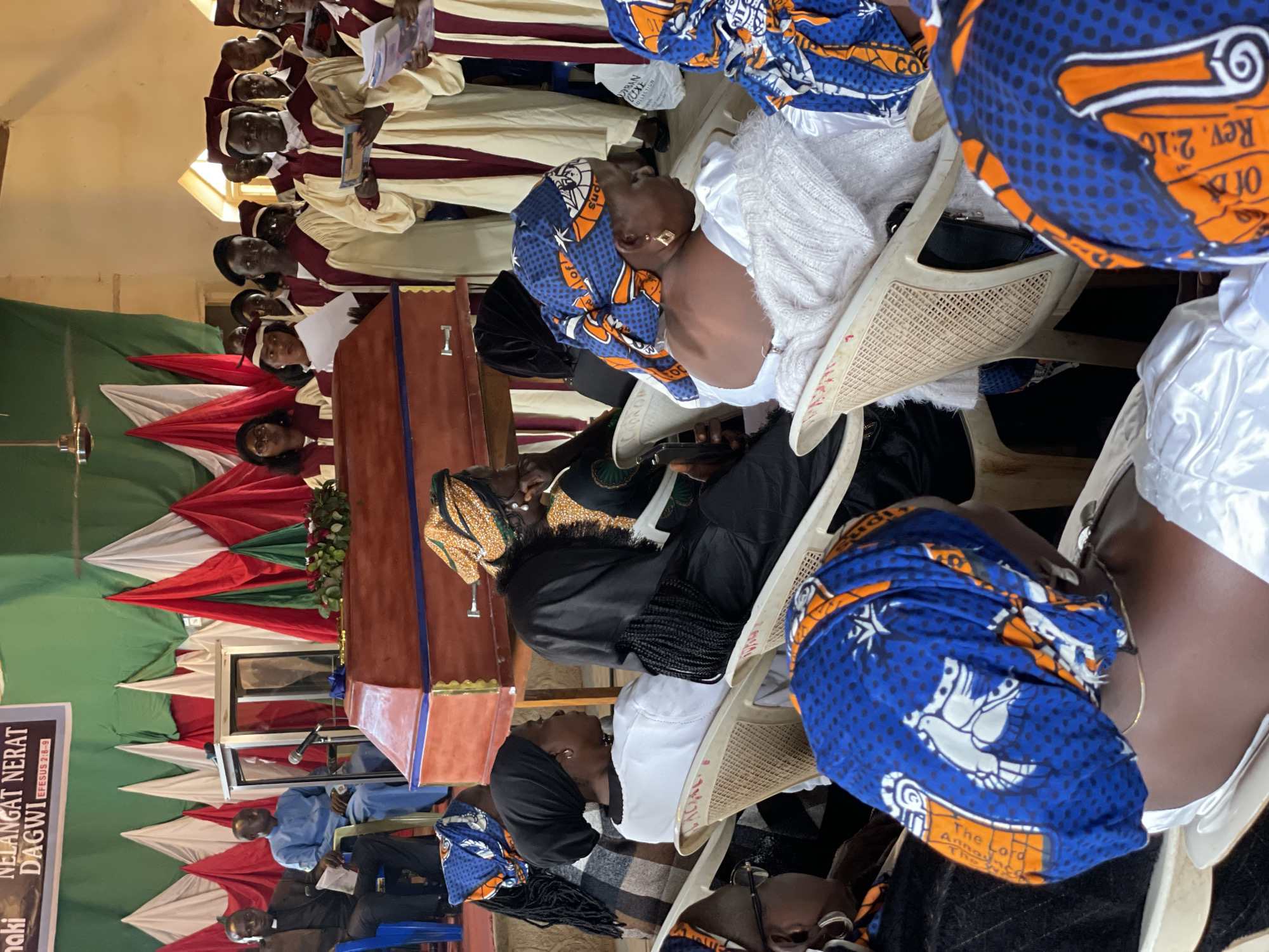 In a building that appears to be a church hall in Kassa, Nigeria, a funeral service is underway. The scene is dominated by a red wooden casket adorned with gold accents, placed centrally on a low wooden stand. A bouquet of red roses rests on the casket.
Several individuals are visible, both seated and standing. To the left of the casket, two men are seated. One, wearing a black clerical collar and robe, holds papers in his lap. The other, in a light blue garment, sits beside him. Behind the casket, a group of people in formal robes, possibly a choir or religious officials, stand and appear to be engaged in the service. They wear cream-colored robes with maroon trim and matching caps, holding papers or books.
The audience consists of individuals, primarily women, seated in rows of plastic chairs. Many are wearing brightly colored headscarves with geometric patterns in blue, orange, and white. Some are dressed in black, while others wear white or cream-colored clothing. The walls of the hall are decorated with draped fabric in red, white, and green, forming a backdrop. A banner with text, including "NELANGAT NERAT DAGWI EFESUS 2:8-9," is visible at the top of the frame. The lighting is a mix of natural light coming from above and possibly interior lights. The overall atmosphere suggests a solemn occasion.