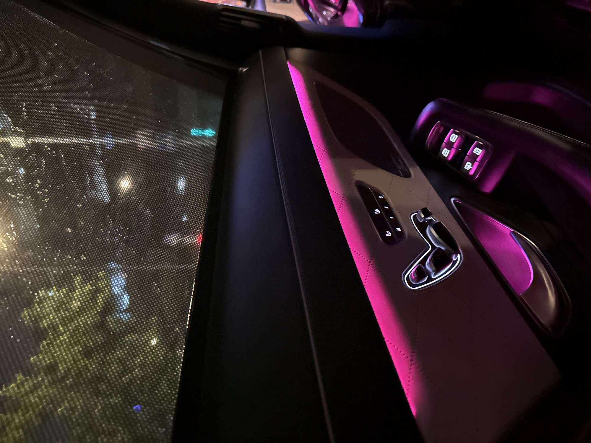 Captured at night, the image features the interior of a car. The scene is dimly lit with pink interior ambient lighting highlighting the door panel, which includes controls for seat adjustments, window controls, and a door handle. Outside the car window, blurred lights and greenery are visible, suggesting an urban environment. While the exact location in Moscow, Russia cannot be determined from the image alone, the nighttime urban setting with possible streetlights and trees suggests a cityscape.