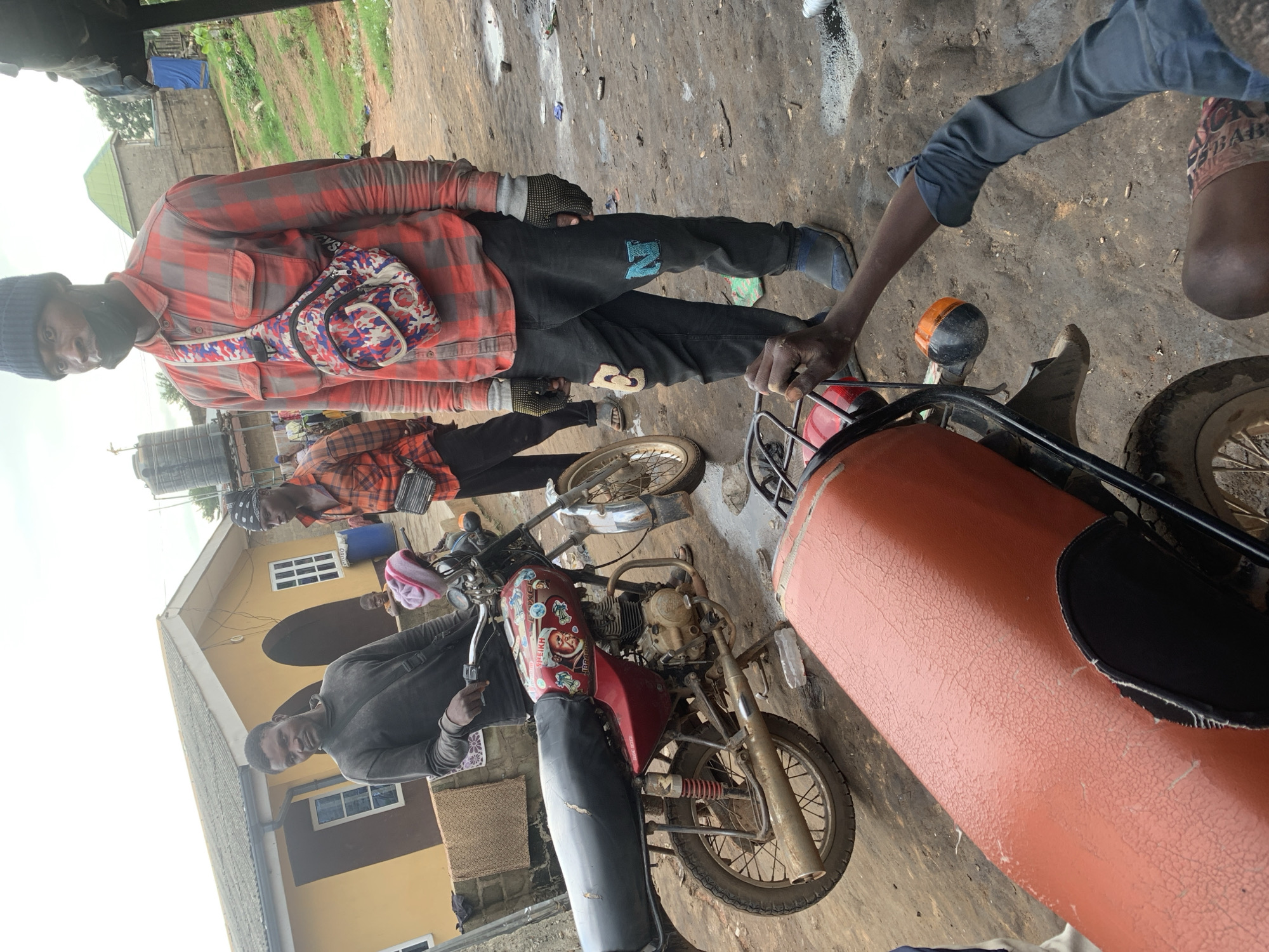 The image depicts an outdoor scene in Shagamu, Nigeria, characterized by a dusty, unpaved ground with scattered debris. In the foreground, an orange motorcycle seat is prominently visible, showing signs of wear and tear with cracked upholstery. To the left of this seat, a partially visible red motorcycle with black seating and a silver exhaust pipe is present. The motorcycle's fuel tank is adorned with stickers, including one featuring a man's face and text.
Three men are the central human subjects. One man, wearing a dark long-sleeved shirt and a black beanie, stands to the left, holding the handlebars of the red motorcycle. Another man, dressed in a red and grey plaid shirt, black pants, grey gloves, and a black face mask, is positioned to the right, wearing a colorful crossbody bag. He appears to be looking towards the camera. Behind these two men, a third man in a similar red and black plaid shirt and a black bandana is also visible.
In the background, a yellow building with brown accents and arched doorways stands. Windows are visible on the building, and a blue barrel is placed near it. A water tank is also visible on a structure behind the building. The overall environment suggests a semi-urban or rural setting, possibly a workshop or residential area. The lighting indicates daylight conditions.