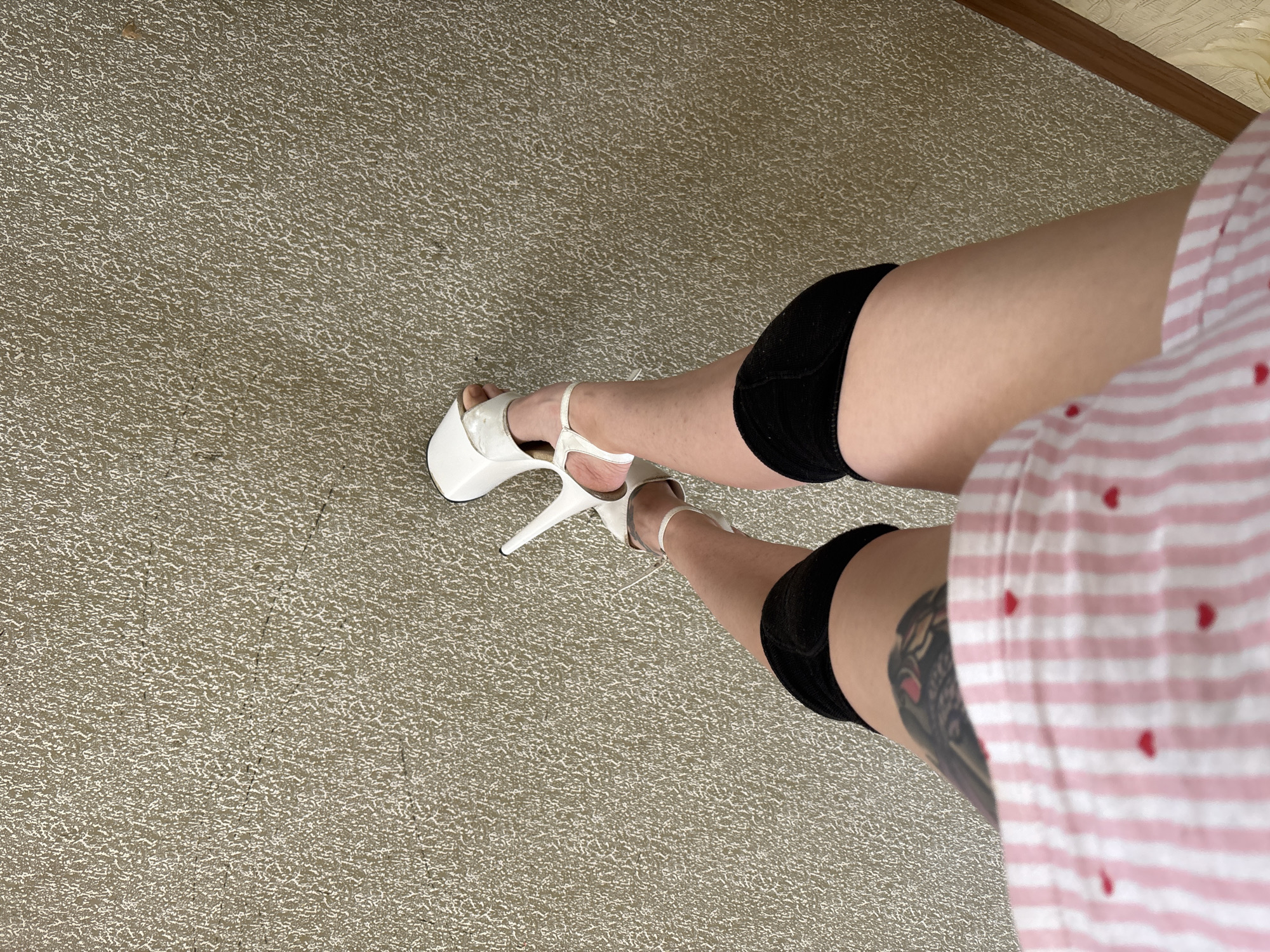 The image shows a person's legs and feet, wearing white platform heels and black knee pads. They are also wearing a pink and white striped shirt with small red hearts. The background is a textured, light-colored wall. There is no indication of the location within Naberezhnye Chelny, Russia.