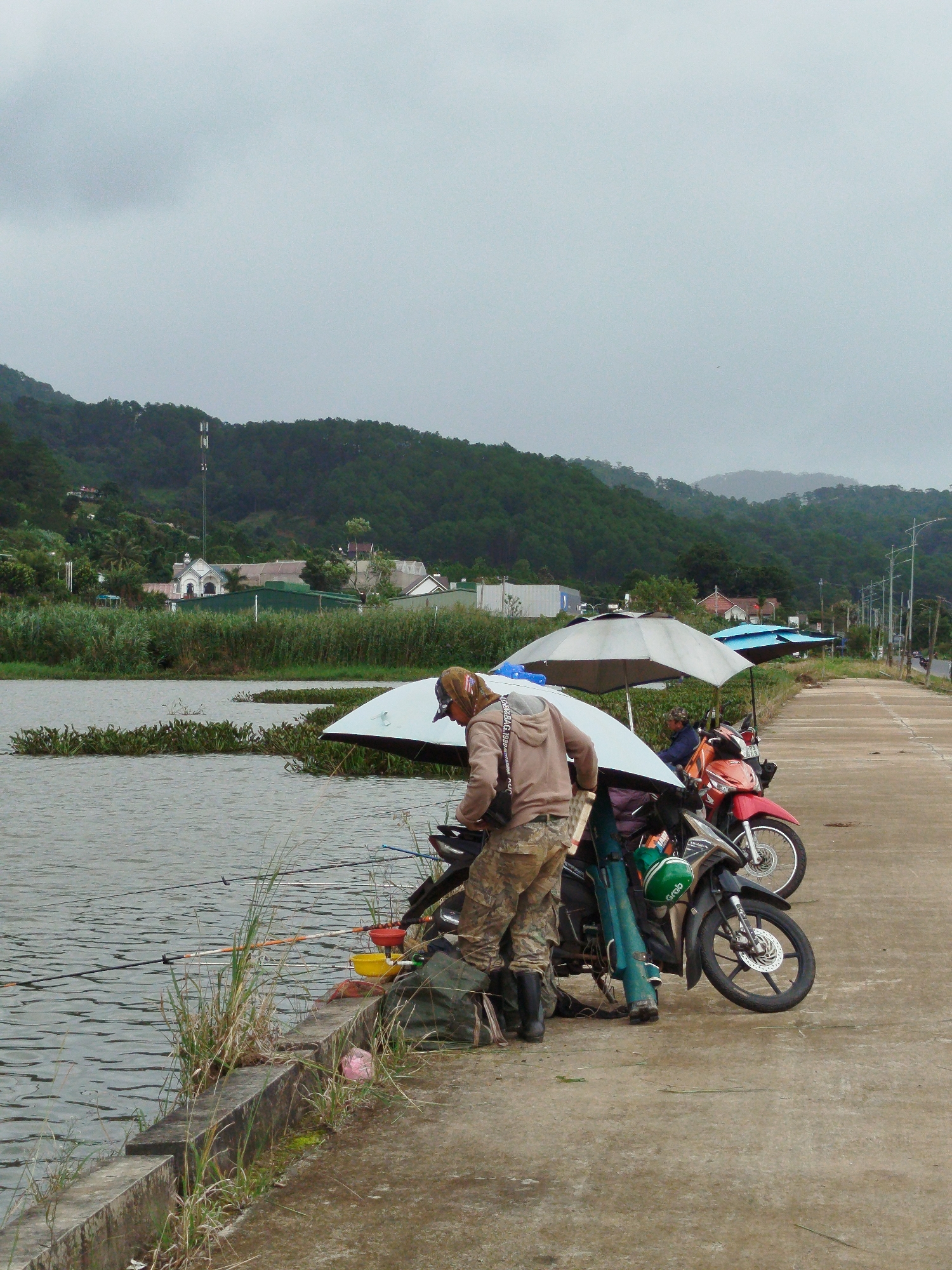 The image depicts a cloudy day by a body of water, likely a lake or river, with a paved road and a hillside covered in dense green trees in the background. The location is identified as K'Long, Vietnam.
In the foreground, a man, wearing camouflage pants and a light brown hooded jacket, is standing next to a motorcycle. He appears to be organizing fishing gear, with fishing rods visible and leaning against the motorcycle. Two umbrellas are set up near the motorcycles, providing shade. One umbrella is white and covers a portion of the motorcycle and the man. Another umbrella, blue in color, is set up behind it.
The motorcycles are parked on the side of the paved road, which runs parallel to the water's edge. The water has some aquatic plants growing in it, and several fishing lines are cast into the water from the bank. On the concrete bank, there are at least three buckets of various colors, including yellow and red, and a pink object, possibly a ball, lies on the ground. Another person is partially visible sitting on one of the motorcycles, wearing a dark top.
The background shows a small settlement with buildings and a prominent white building with a red roof, possibly a church or a significant public building. A tall telecommunications tower is visible on the hillside. The overall atmosphere suggests a quiet, possibly rural setting, with the people engaged in recreational fishing.