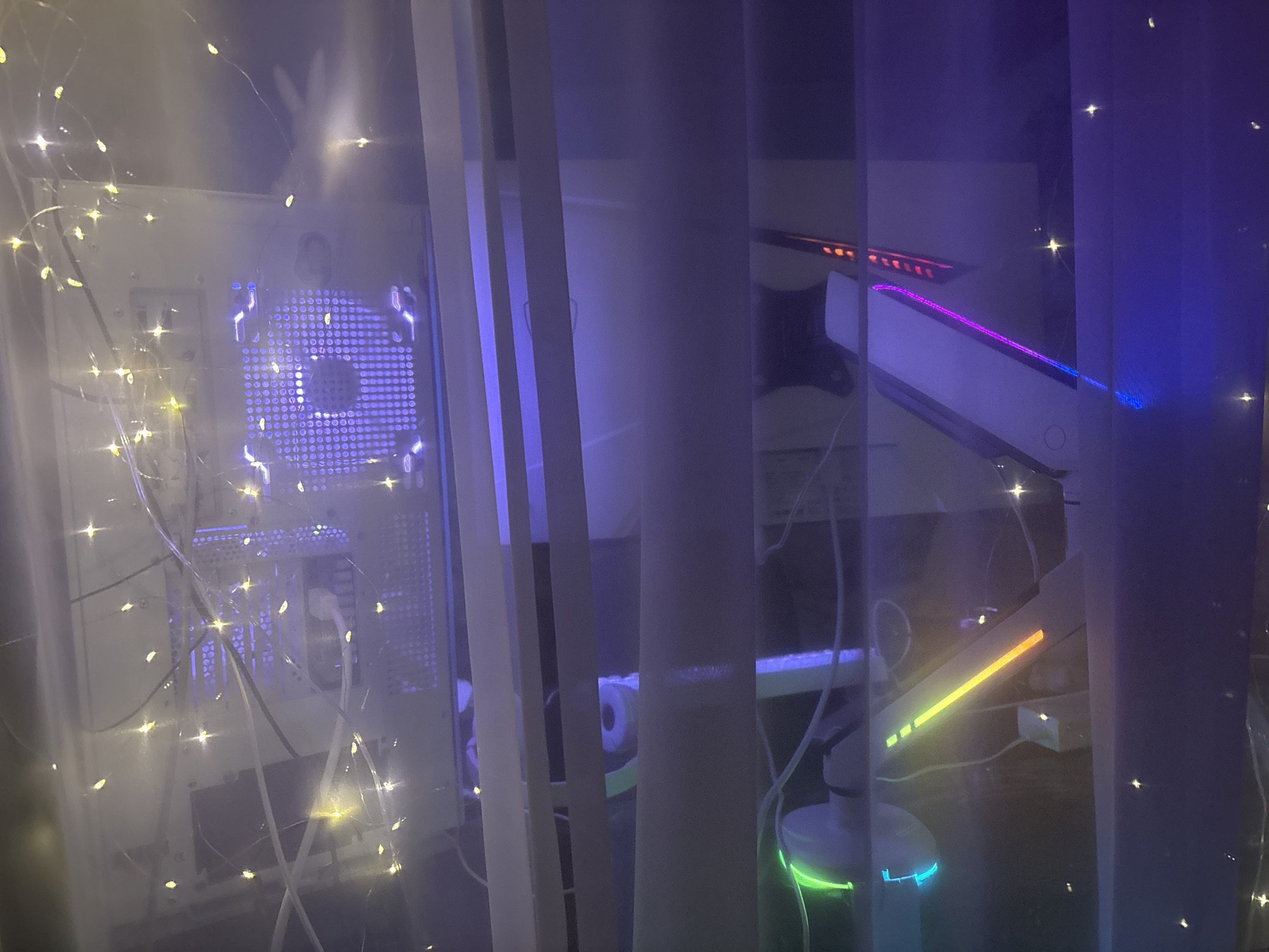 This media file captures a dimly lit interior scene, likely a room, viewed through a window with sheer curtains. The room appears to be a gamer's setup, featuring a white computer tower with glowing purple lights emanating from within, possibly from internal fans or RGB lighting. The computer tower is adorned with a string of small, warm-toned fairy lights, creating a festive and somewhat cluttered aesthetic.
Adjacent to the computer, another electronic device is visible, possibly a monitor or a display, which also exhibits colorful RGB lighting, shifting from pink to blue and yellow. A gaming keyboard with illuminated keys can be seen, as well as a portion of a gaming mouse with green and blue lighting. The overall impression is of a technologically-focused space designed for entertainment, with an emphasis on ambient lighting.
The presence of the fairy lights and the purple and pink hues from the computer components suggest the scene might be captured during the evening or nighttime, enhancing the atmospheric glow of the lights. The sheer curtains, illuminated by the interior lighting, create a diffused effect, partially obscuring the view and adding a layer of mystery or privacy to the scene. There is no indication of people, specific weather conditions, or any discernable text in the image. The setting appears to be indoors, possibly a bedroom or a dedicated gaming area.