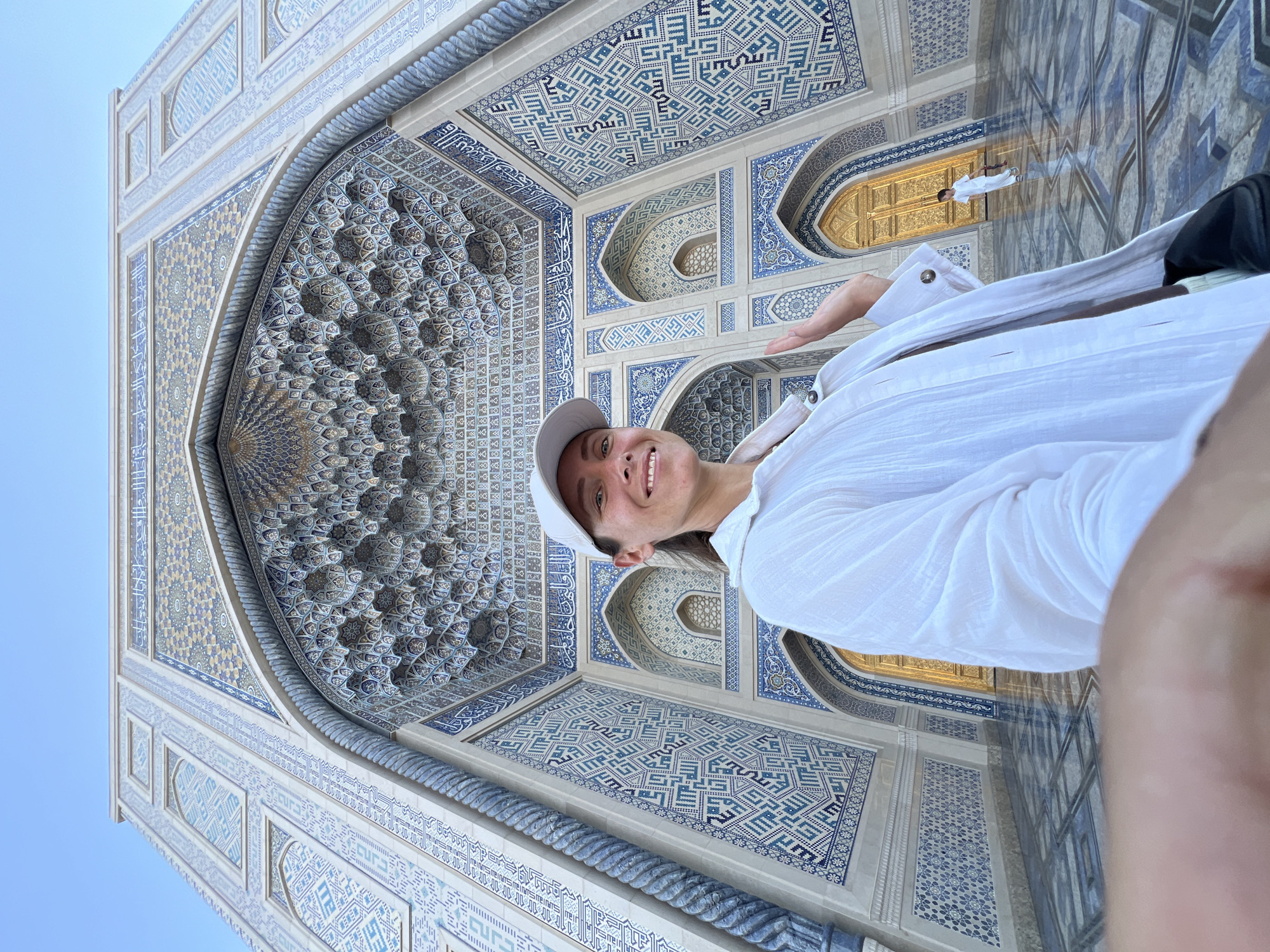 The image is a selfie taken by a person in front of an elaborately decorated building with intricate geometric tilework in shades of blue, white, and gold. The architecture features arches, recessed niches, and detailed patterns. The subject is wearing a white shirt and a white baseball cap, smiling at the camera. Their right hand is extended, pointing towards the building.
In the background, through a golden doorway, another person dressed in white can be seen standing. The reflective surface of the ground creates a mirror image of the architecture and the person in the doorway.
The setting appears to be an outdoor location with a clear blue sky visible at the top left of the image. The style of the architecture, particularly the geometric patterns and the use of blue and white tiles, suggests a location in Central Asia, specifically Tashkent, Uzbekistan, known for its historical and Islamic architecture. The lighting indicates it is daytime. The overall mood is positive and enthusiastic, conveyed by the subject's smile. There is no visible text in the image.