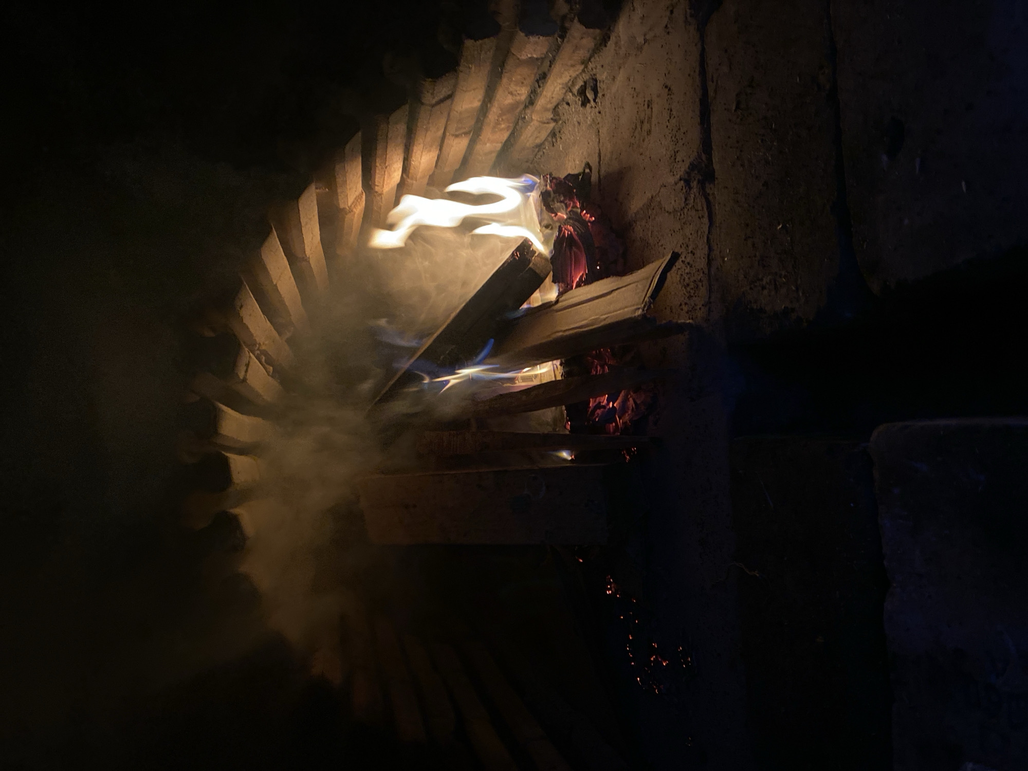 The image shows a fire burning in an indoor fireplace. Flames and smoke are visible, along with burning wood. The fireplace is constructed with brick and concrete and is in the city of Konstantinovo, Bulgaria.