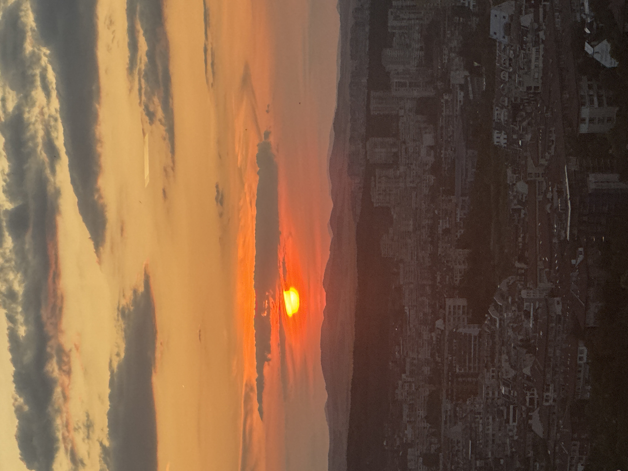 This image depicts a sunset over the city of Sofia, Bulgaria. The sun, a bright orange-yellow orb, is partially obscured by a strip of dark cloud, casting a warm glow across the sky. The sky transitions from vibrant orange near the horizon to a softer yellow and pink higher up, with scattered grey clouds adding texture. In the middle ground, a range of low, hazy mountains stretches across the landscape. Below the mountains, the city of Sofia unfolds, a dense collection of buildings with varying rooflines and architectural styles. Apartment buildings and smaller residential structures are visible, appearing in shades of grey and brown due to the dimming light. The overall impression is of a tranquil urban evening, with the natural beauty of the sunset juxtaposed against the man-made environment of the city.