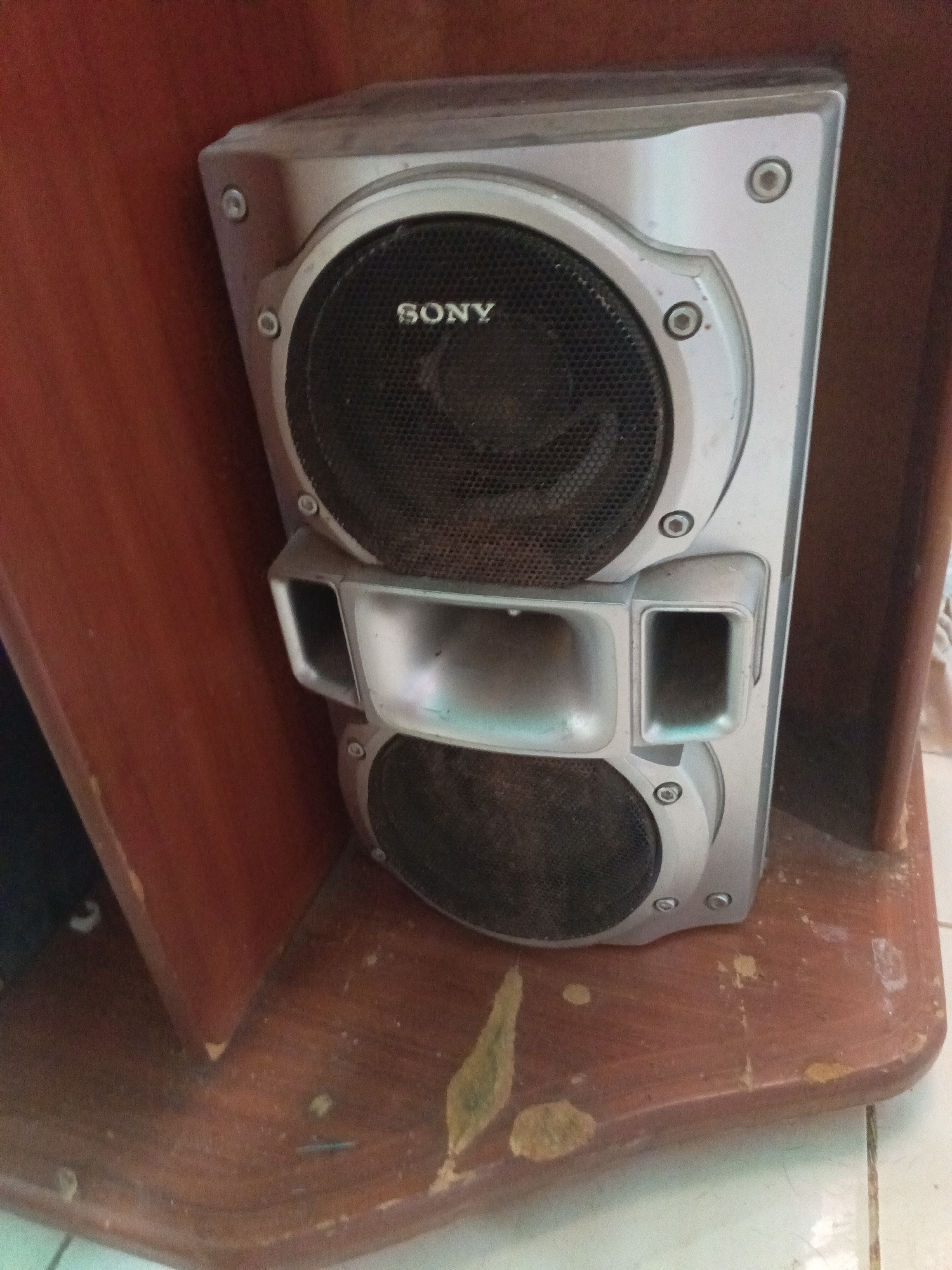 An aged Sony speaker stands on a worn wooden shelf, nestled against a matching wooden wall or cabinet side. The speaker, primarily silver-grey, features two prominent black mesh grilles, with the "SONY" logo clearly visible on the upper grille. Between the two grilles are two rectangular, silver-grey ports or vents, adding to its functional design. Multiple small screws or fasteners are visible on the speaker's casing, securing its components.
The entire unit, particularly the speaker grilles and casing, is covered in a layer of dust, suggesting it has been in its current position for some time without cleaning. The wooden shelf below the speaker exhibits significant wear and tear, with numerous chips, scratches, and peeling sections revealing a lighter wood or undercoat. Dust and small particles are also scattered across this wooden surface. A similar wooden panel forms the backdrop to the left, while the bottom right corner shows a glimpse of white or light-colored floor tiles with dark grout lines. The scene depicts an indoor setting, likely a home, showcasing an older, well-used audio component within its environment. No human activity is depicted.