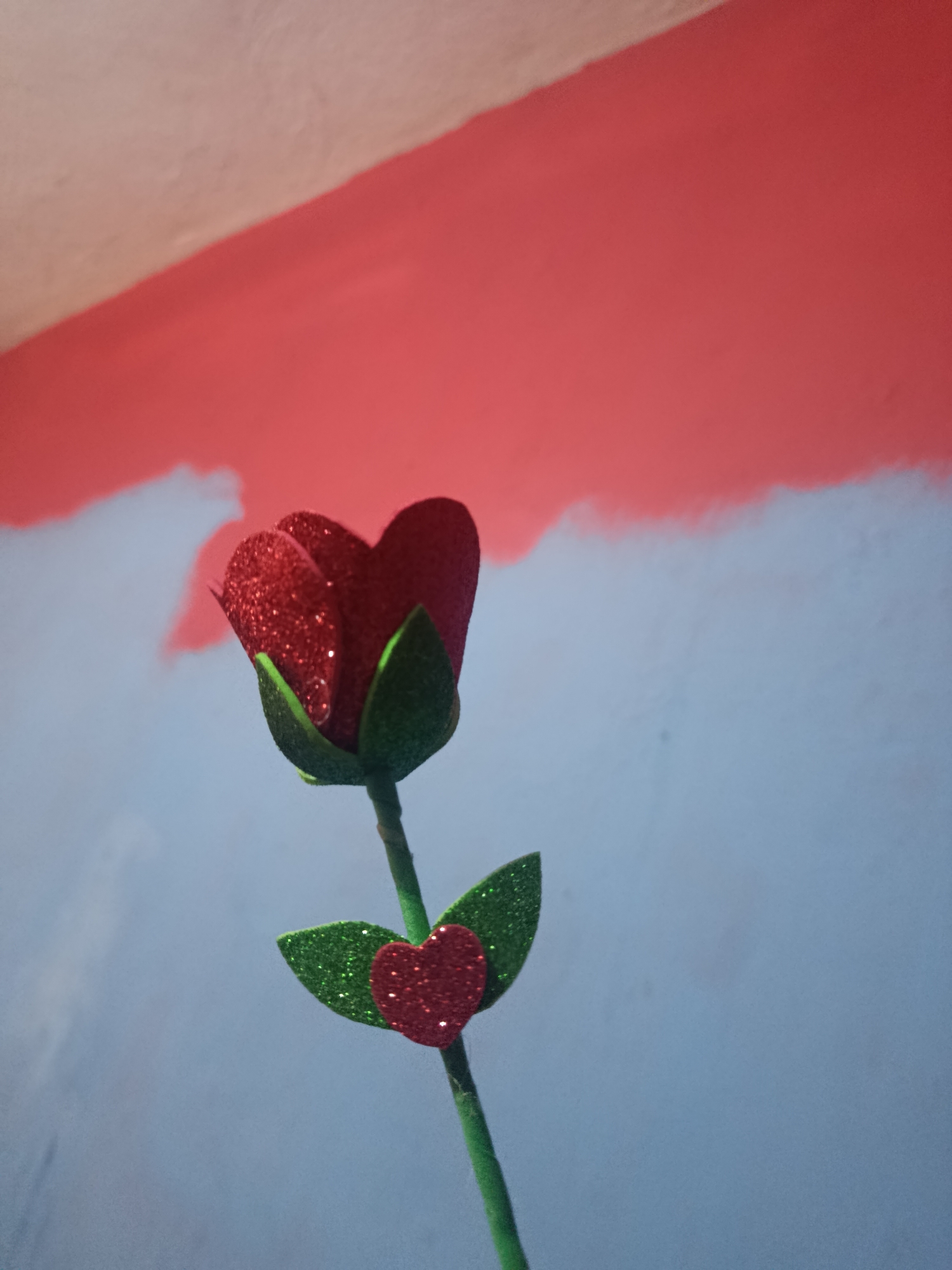 A close-up shot of a single, artificial rose against a painted wall. The rose has a red, heart-shaped flower made of glittery material, green leaves with glitter, and a green stem. A small, red, glittery heart is attached to one of the leaves. The wall is painted with a broad horizontal stripe of reddish-pink at the top, and a blue-gray color below. The location is in Juan Griego, Venezuela.