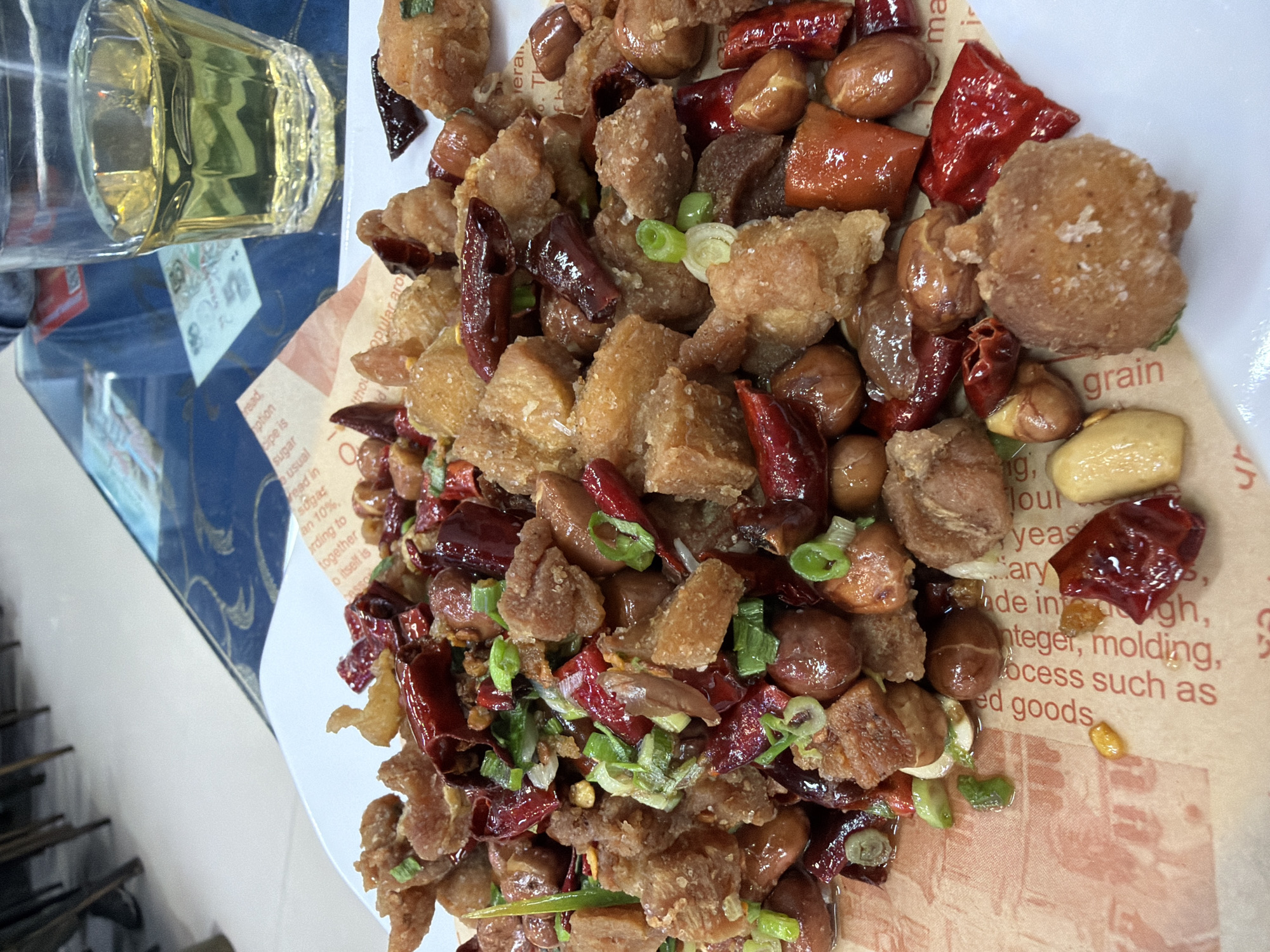 The image captures a plate of spicy, stir-fried food, likely a Chinese dish such as "La Zi Ji" (Sichuan Chili Chicken) or a similar preparation featuring fried meat. The main subjects are numerous small, golden-brown pieces of what appears to be crispy fried chicken or pork, generously mixed with whole roasted peanuts, bright red dried chili peppers (some whole, some broken), and chopped green onions. The food is served on a white plate lined with a piece of brown, textured paper. The chili peppers exhibit a glossy, oily coating.
To the upper left of the food, a clear glass filled with a light yellow liquid, possibly an alcoholic beverage like baijiu or a type of tea, stands on a dark blue surface. The background is blurred but suggests an indoor setting, possibly a dining table. On the far left, a metallic structure resembling a rack or shelves is partially visible.
The activity depicted is a meal. The paper under the food contains legible English text, including fragments such as "read," "popular aro," "ption," "recipe is," "sugar," "is usual," "ed in," "an 10%," "according to," "together," "o itself is," "grain," "integer, molding," "process such as," and "ed goods." No people are visible, and the time of day or weather cannot be determined from the image.