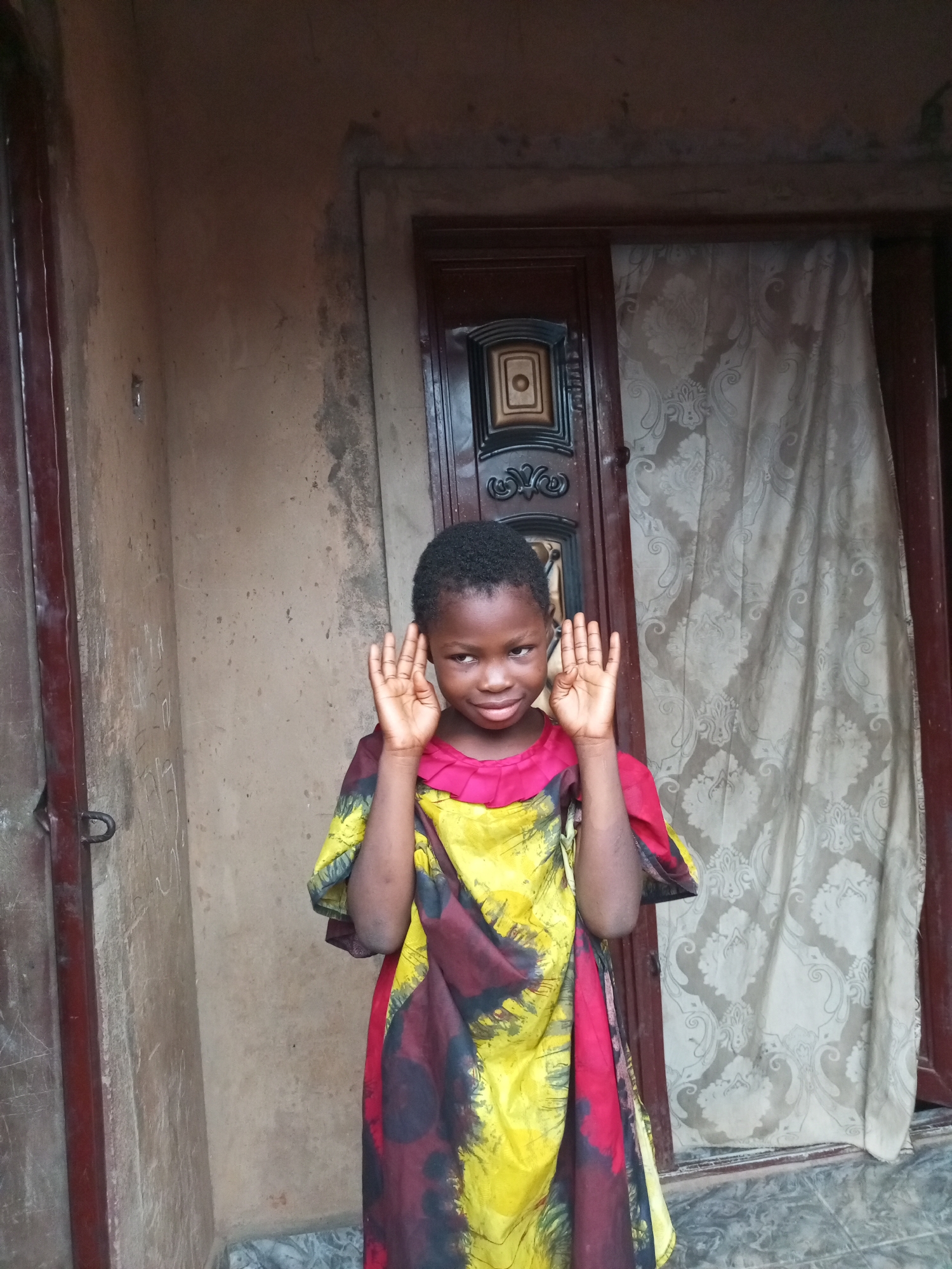 A young girl stands in front of a doorway in Tempure, Nigeria. She is wearing a colorful dress with yellow, red, and black patterns. Her hands are raised to her face, with her fingers spread apart. The wall behind her is made of stucco and shows signs of wear and tear. The doorway has a dark wooden frame and a decorative panel in the center. A patterned curtain hangs on the right side of the doorway. The ground in front of the doorway is tiled.